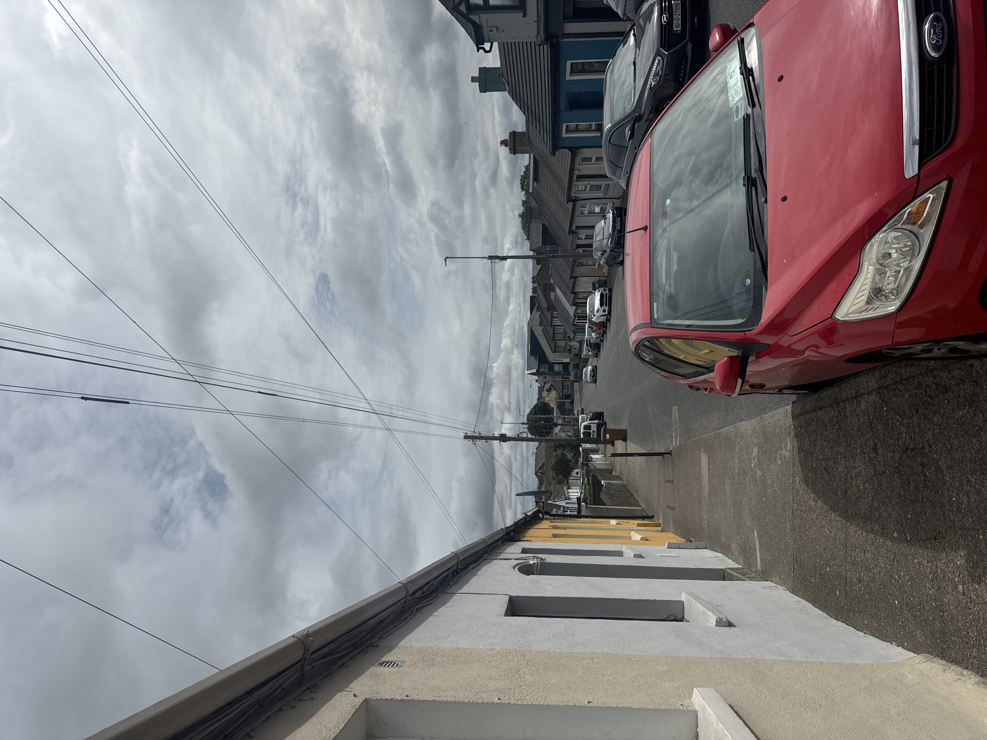 A street scene is depicted in Waterford, Ireland, under an overcast sky. The perspective is from the sidewalk, looking down a residential street lined with houses on both sides. A red Ford Focus is parked in the foreground on the right, partially obscuring the view of the street. The houses are of varying colors, including white, yellow, and blue, with pitched roofs. Power lines crisscross the sky, supported by utility poles. A few other cars are visible parked along the street and driving in the distance. The sidewalk is made of concrete, and a low wall separates the properties from the street. The overall impression is a typical urban street scene in a temperate climate.