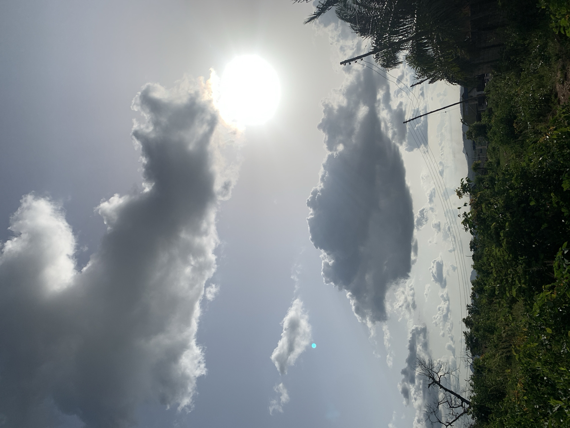 The image, taken from a slightly low angle, shows a partly cloudy sky with the sun prominently visible. Two large cloud formations dominate the left and center of the frame. The left cloud is lighter, while the central one is darker and denser. Smaller, scattered clouds appear in the sky.
In the background, on the right side of the image, dense green vegetation and trees are visible. A few buildings can be discerned amongst the foliage. Power lines stretch across the sky, intersecting with the trees.
The location is identified as Gbeleko, Nigeria. There are no people visible in the image.
