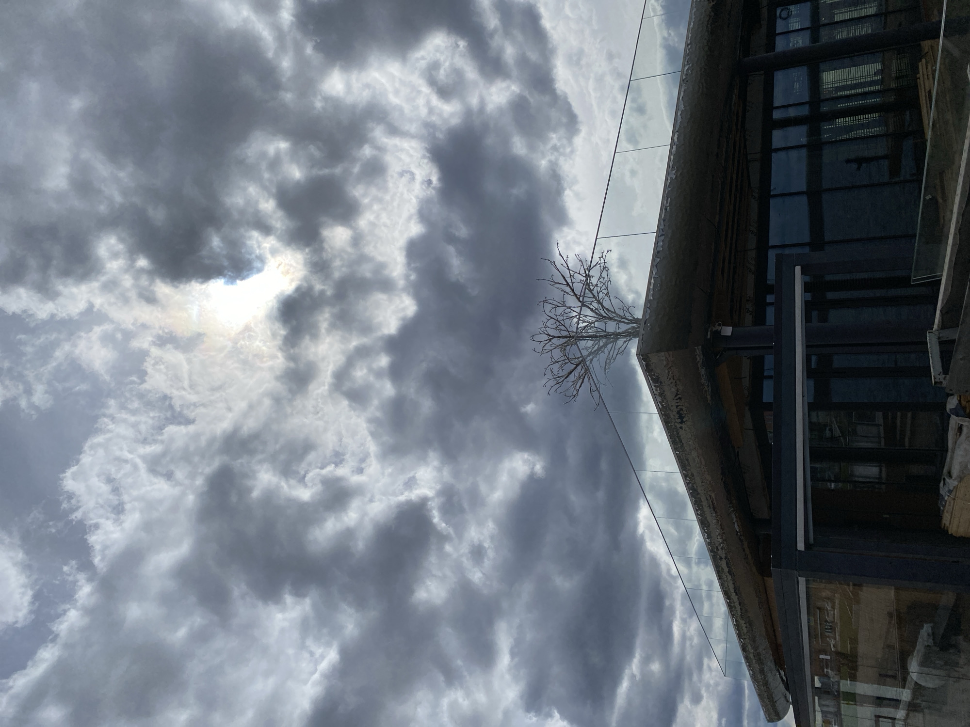 The image shows a cloudy sky with a small patch of sunlight breaking through. A modern building with glass panels is visible on the right side of the frame. A bare tree stands near the corner of the building. The location is likely Votkinsk, Russia, though no specific landmarks or features are identifiable to confirm this. No people, actions, or interactions are visible. The environment is cloudy and the building appears to be situated in an urban area.
