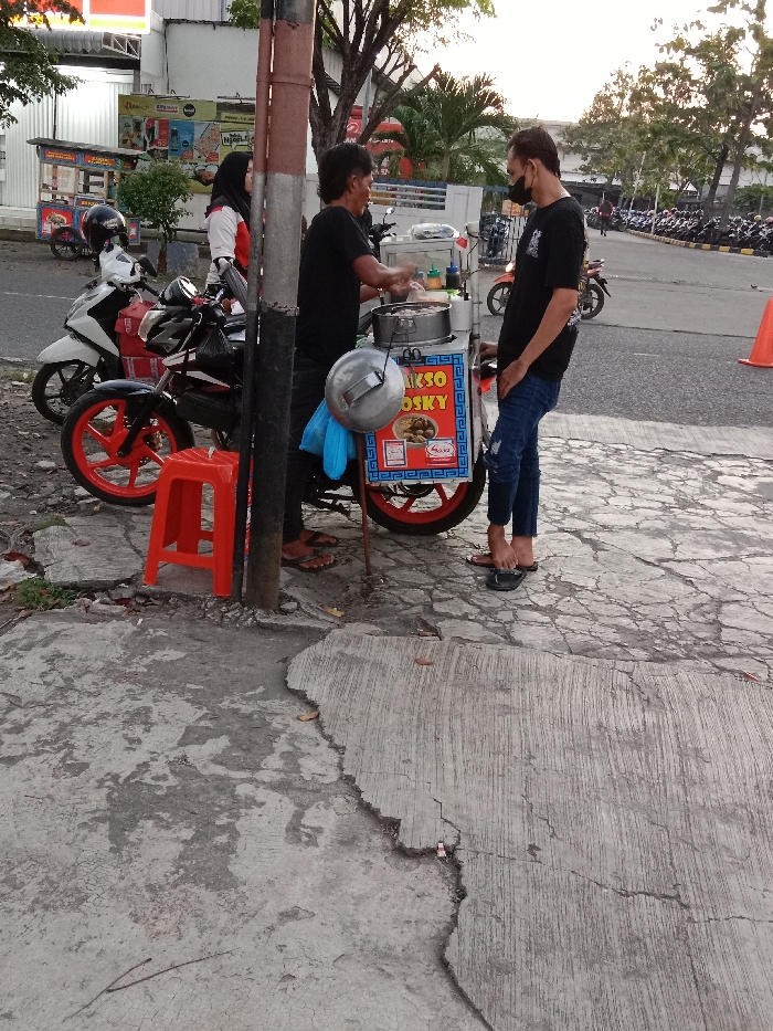 The scene takes place on a cracked concrete sidewalk in front of a street food vendor in Tugu, Indonesia (latitude: -6.973, longitude: 110.326). A man wearing a black shirt and blue jeans is standing in front of the vendor, interacting with a man wearing a blue shirt. There is a red stool next to the food vendor, which appears to be a motorcycle with a sidecar. The vendor is selling food from a large, metal cart with red and blue lettering. There are a couple of other people in the background but their faces are not visible. There is a motorcycle to the left of the vendor, and another in the background, along with parked cars. There is a large tree to the left of the food vendor and a lamppost behind it. The vendor is holding a container filled with food ingredients while the man in the blue shirt holds a small white container. The vendor has a silver helmet at his feet. The sidewalk is grey and cracked. The sky is blue with a light cloud cover. The image is taken at a slightly low angle, looking up toward the vendor.