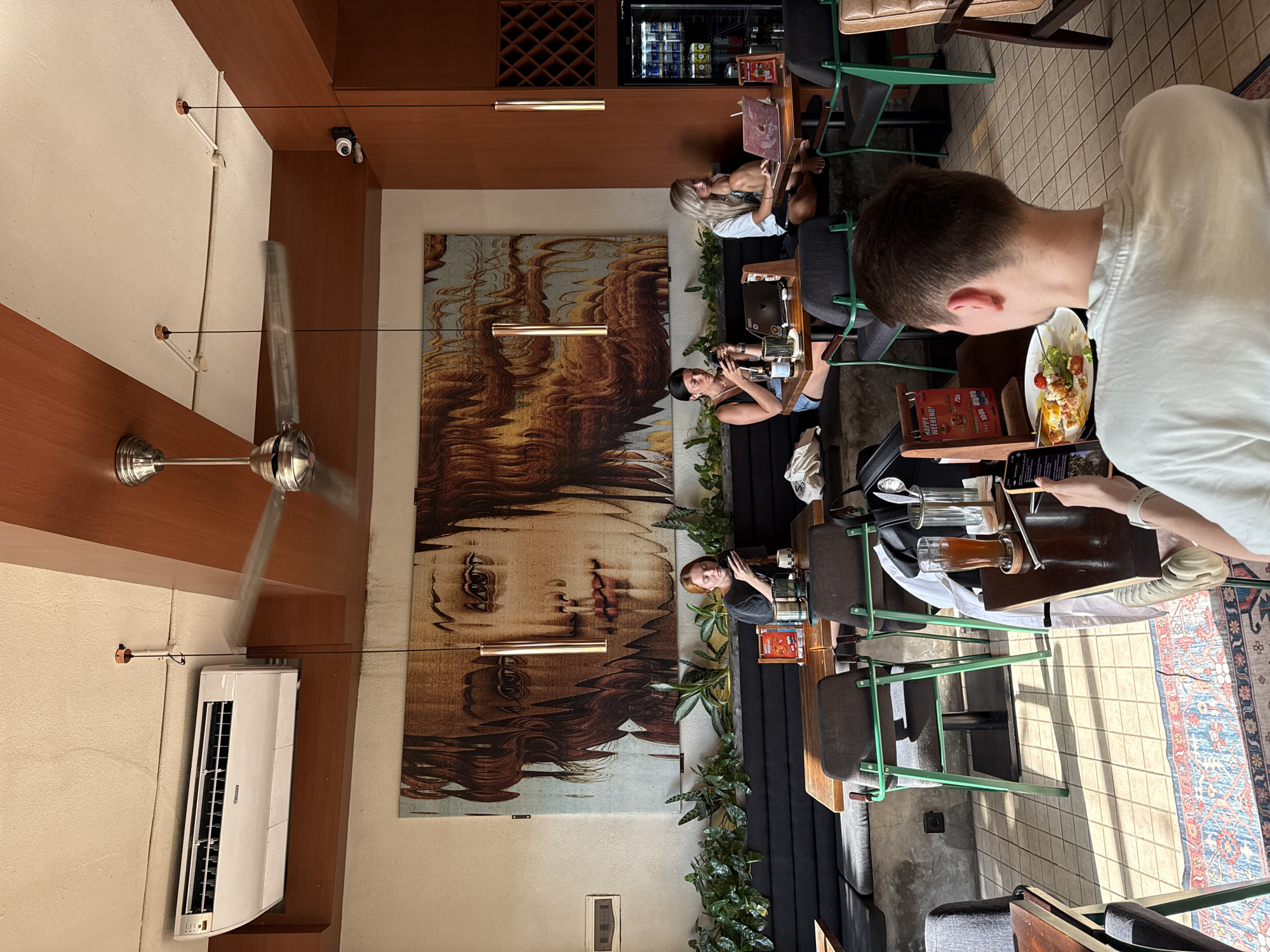 The image shows the interior of a restaurant or cafe in Uluwatu, Indonesia. Several people are seated at tables, appearing to be customers. Some are using laptops and phones, while others have food and drinks in front of them. The establishment features an eye-catching mural of a woman's face on the wall. The room is lit by pendant lights and has an air conditioner unit mounted on the wall. The dining area is decorated with greenery, and the decor includes a rug. The cafe setting suggests a relaxed, modern atmosphere.