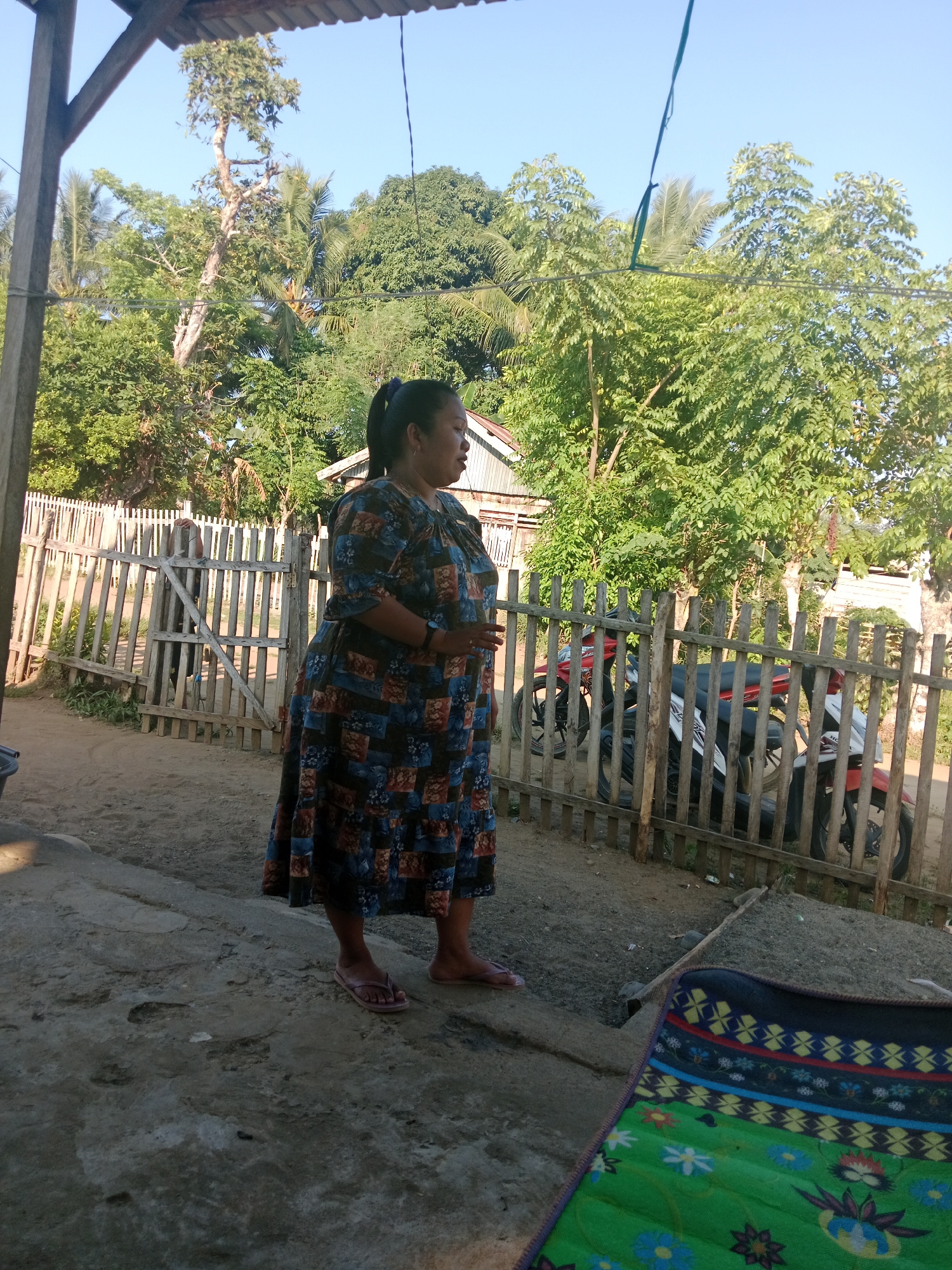 A medium-shot, eye-level, outdoor, daytime photograph captures a woman standing on a concrete surface next to a dirt path in a rural area of Lakea Satu, Indonesia. The woman, who appears to be of medium build and ethnicity, is wearing a long, patterned dress with a predominantly blue and orange color scheme, and flip-flops. She is looking to her right, her left hand is resting on her stomach, and her right hand is down by her side. Behind her is a weathered wooden fence, through which two motorcycles are visible: a red one and a white and red one. The fence encloses an area with more trees and vegetation, and beyond that, a building with a white roof and a light-colored exterior. To the left of the woman, a wooden post and part of a roof structure are visible, suggesting she is standing under an overhang. The ground consists of both concrete and sandy soil. In the foreground, to the right, a portion of a colorful mat with a floral pattern is visible. The sky is clear and blue.