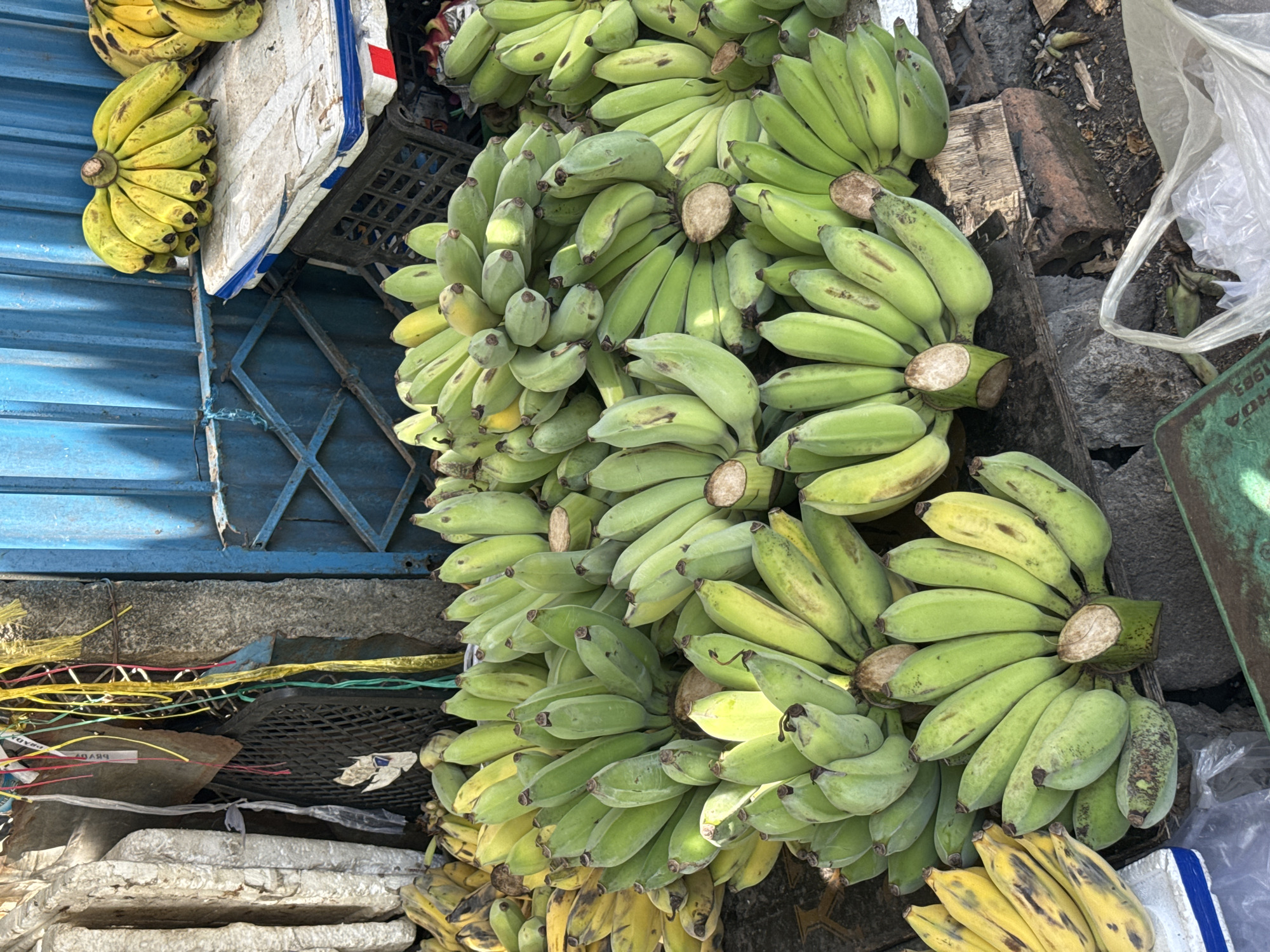 The image shows a close-up of a street market stall selling numerous bunches of green and yellow bananas. The bananas are arranged in various piles on the ground and on a white flat surface. The background consists of a blue corrugated metal wall with some metal grating and various pieces of debris, including what appear to be old white blocks or mats and some trash. Colorful plastic strings are hanging from above. The ground is dirt and littered with small stones and debris. A plastic bag is visible in the lower right corner of the image. The overall impression is that of a typical outdoor market in Vietnam, likely in Nha Trang Ward, given the context. There are no people visible in the image.