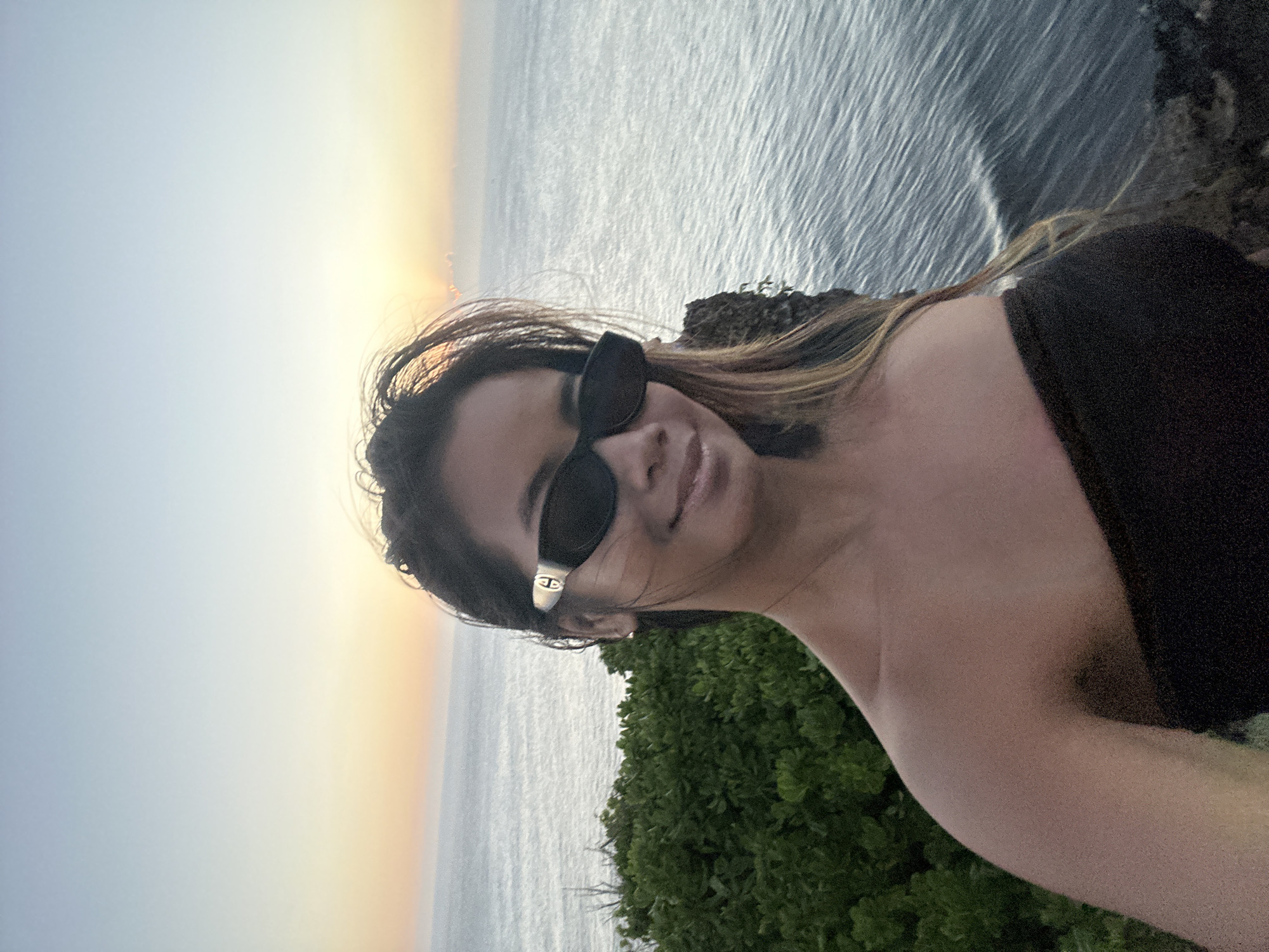 A woman with dark hair, wearing black sunglasses and a black strapless top, takes a selfie on a cliff overlooking the ocean. The sun is setting, casting a warm glow across the sky and water. Lush green bushes are visible on the left side of the image. The location is the coast of Kuta Selatan, Indonesia.