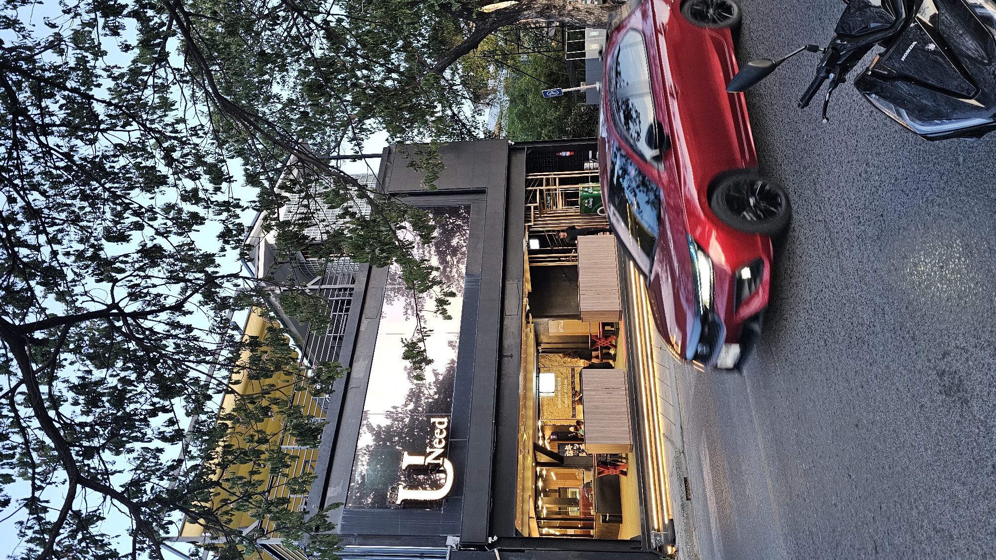 This is a slightly angled, eye-level shot of a street scene in Baan Chaweng, Thailand. A bright red car is parked on the right side of the road, and a black motorcycle is parked behind it. The car's headlights are on, and the motorcycle has the brand name "HONDA" visible on its front.
The building on the left appears to be a commercial establishment with large windows. Through one of the windows, the text "D Need" is visible, suggesting the name of the establishment. The interior of the building is lit and seems to be a dining or seating area with tables and chairs. The overall atmosphere suggests it's late afternoon or early evening, as the sky is a soft blue with hints of sunset colors reflecting in the building's windows. Trees with lush green foliage frame the scene, with branches extending into the frame from the top left. The road is asphalt, and the lighting suggests natural light mixed with artificial illumination from the building.