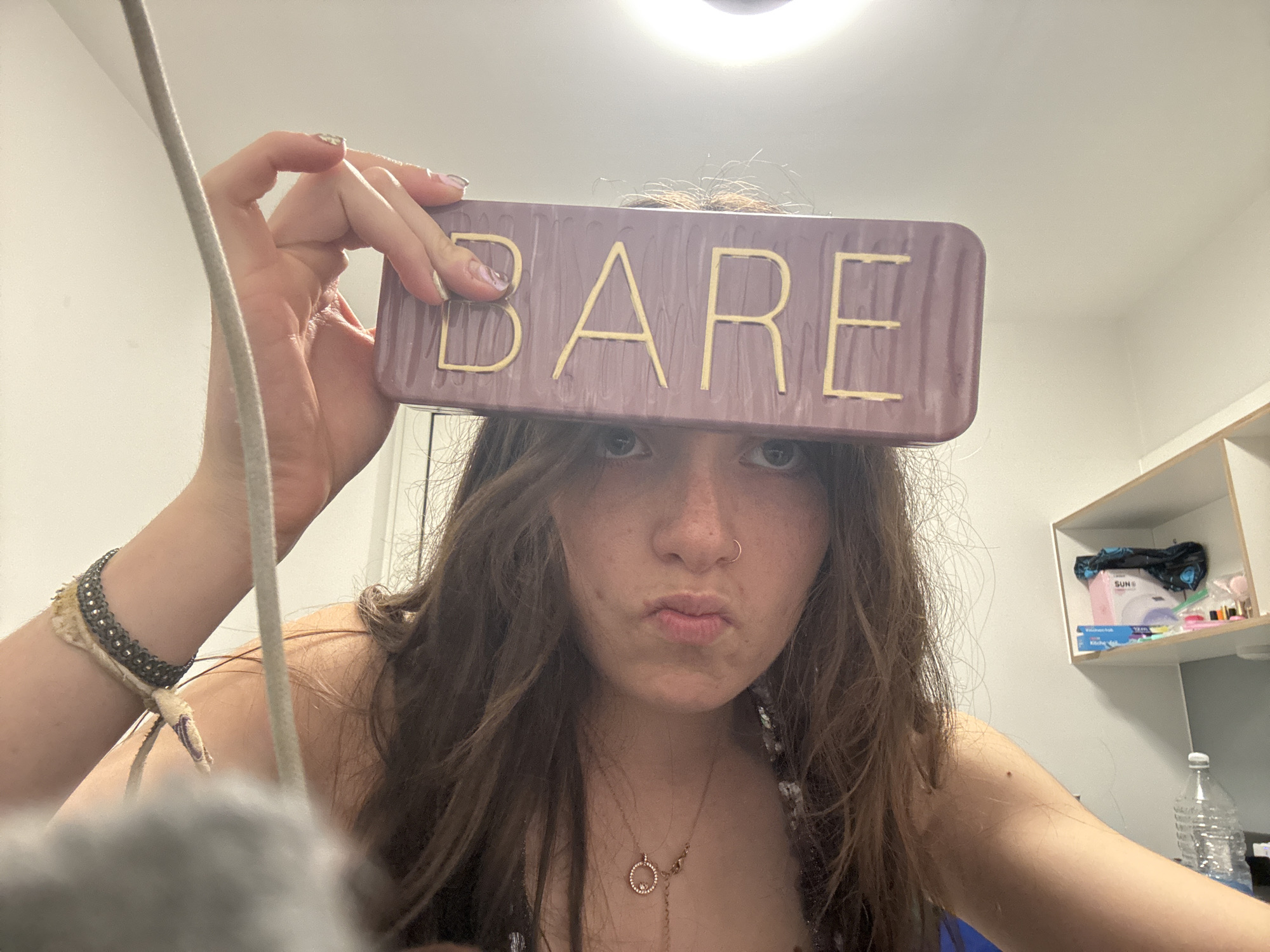 A young individual, likely female, with long, wavy medium-brown hair, poses for a close-up photograph indoors. She holds a rectangular, reddish-purple object with the word "BARE" written in gold capital letters across her forehead, obscuring the upper part of her face except for her eyes. Her lips are pursed in a pout, and she has a small nose ring on her left nostril.
She is wearing a delicate chain necklace with a circular pendant containing small stones. On her left wrist, she has a multi-layered bracelet made of fabric and metal beads. Her nails are manicured with a light, possibly iridescent, polish.
The setting appears to be a simple indoor room, possibly a bedroom or dorm, indicated by plain white walls and a white shelving unit in the background on the right. On the shelves, a pink box with the brand "SUN" visible, along with other small containers and a black fabric item, can be seen. Below the shelves, a clear plastic water bottle is partially visible. A light fixture is discernible at the top center of the frame. A grey, fuzzy object and a light-colored cord run vertically along the left side, partially obscuring the scene. The lighting is artificial.