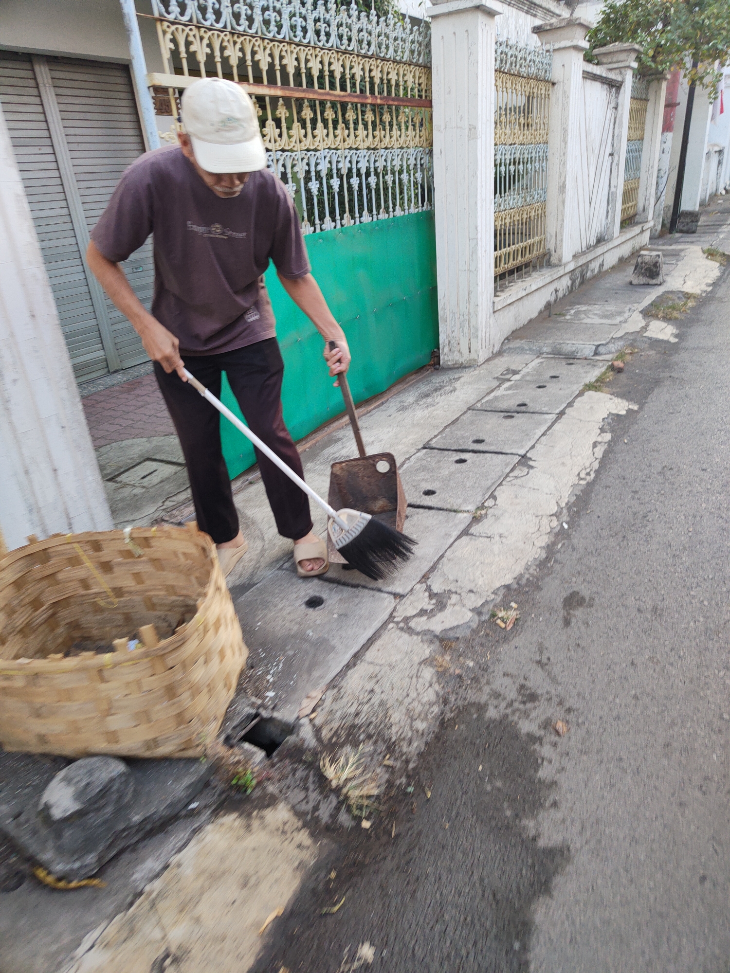 A person is sweeping debris with a broom and dustpan onto the sidewalk in front of a residential building, possibly in Kauman, Indonesia. The person, wearing a brown t-shirt, dark pants, a light-colored cap, and sandals, is standing on a concrete sidewalk adjacent to a paved road. A large woven basket is positioned to the left of the person, presumably to collect the swept debris. The background features a white-patterned fence, a green gate, and a closed metal door. The ground is a mix of concrete sidewalk, paved road, and some patches of dirt and vegetation. Drainage grates are visible on the sidewalk. The lighting suggests it is daytime.