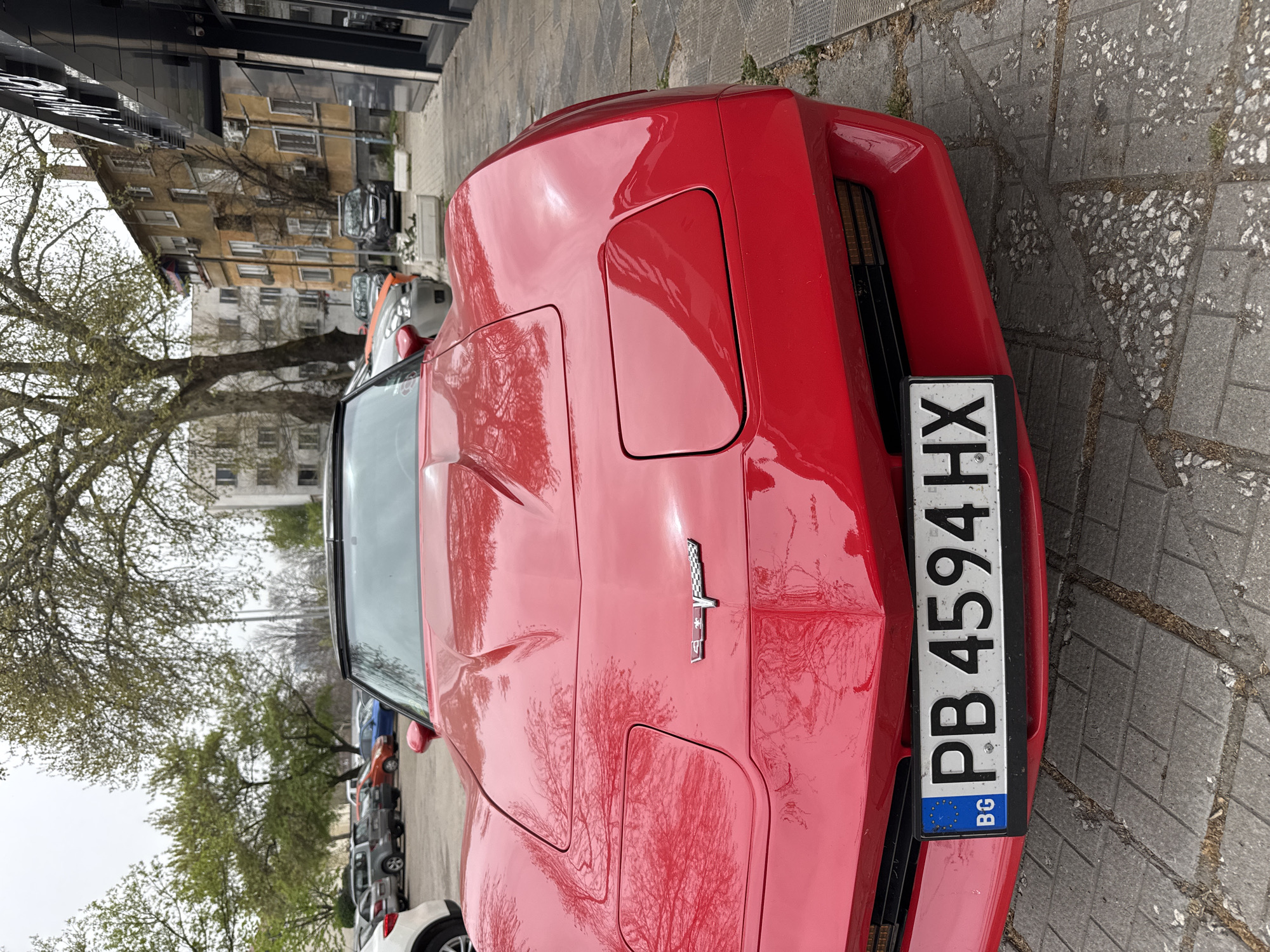 The image shows the rear of a red Chevrolet Corvette parked on a paved sidewalk. The car has a Bulgarian license plate "PB 4594 HX". The street has buildings visible with trees lining the street. The license plate indicates the car is registered in the Plovdiv region. Therefore, the image is from Plovdiv, Bulgaria. Other vehicles are parked nearby. The weather appears overcast.