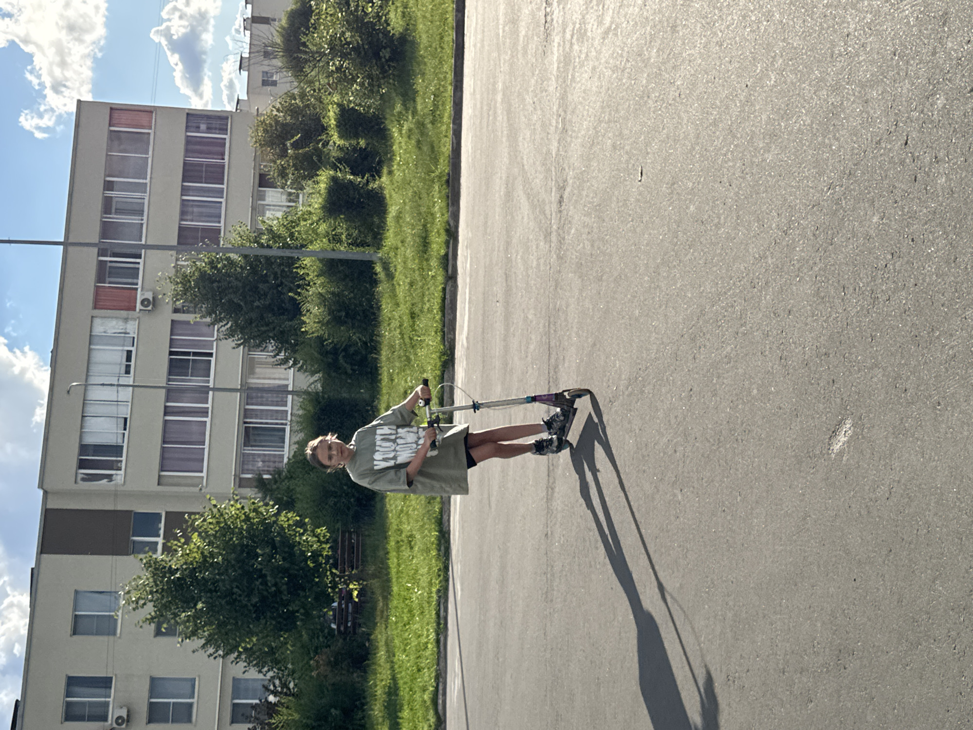 A young girl with blonde hair, wearing glasses and a green oversized t-shirt with a graphic print, rides a scooter on a paved surface. She is facing the viewer and appears to be in motion. The scooter is silver and purple. Her shadow is cast long to the left on the asphalt. Behind her, a strip of green grass borders a row of bushes and trees. Further back, a multi-story apartment building with many windows and balconies is visible. The sky above is blue with scattered white clouds. The scene appears to be an urban residential area, likely in Domodedovo, Russia, given the architectural style of the building and landscaping.