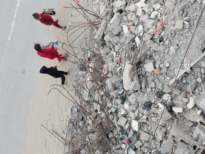 Three children walk from left to right on a sandy strip adjacent to a paved road, overlooking a substantial accumulation of building rubble and debris in Khan Yunis, Palestinian Territory. The child on the left wears a black hooded tracksuit. The middle child wears a red hooded tracksuit and carries a light-colored object. The child on the right also wears a red hooded tracksuit, carrying a black and silver rectangular object. The foreground is dominated by extensive piles of grey concrete fragments, broken blocks, and exposed, twisted rebar, intermingled with various items of trash. The light grey asphalt road occupies the upper portion of the frame. There are no explicit interactions between the children.