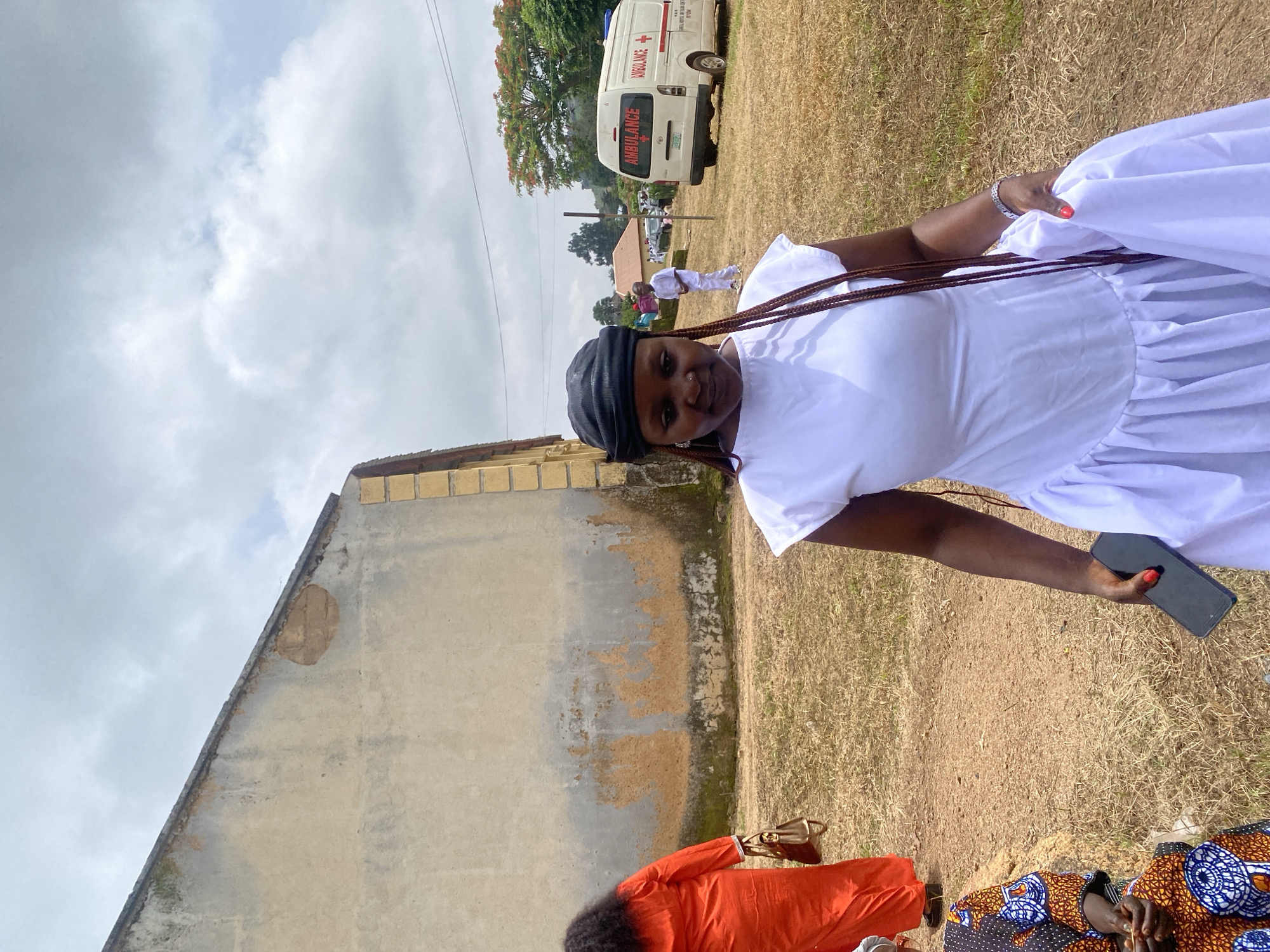 An outdoor scene in the city of Riyom, Nigeria, features a woman in a white dress in the foreground, her gaze directed downward. She wears a black headscarf and her braided hair cascades over her shoulder. In her right hand, she holds a dark smartphone, and a silver wristwatch adorns her left wrist. The background reveals an aged, neutral-toned building with signs of wear and tear. Further back, an ambulance with "AMBULANCE" written on its side is parked, suggesting a medical or emergency context. Other individuals are visible in the distance, some in white attire. The ground is covered in dry grass, and the sky is cloudy.