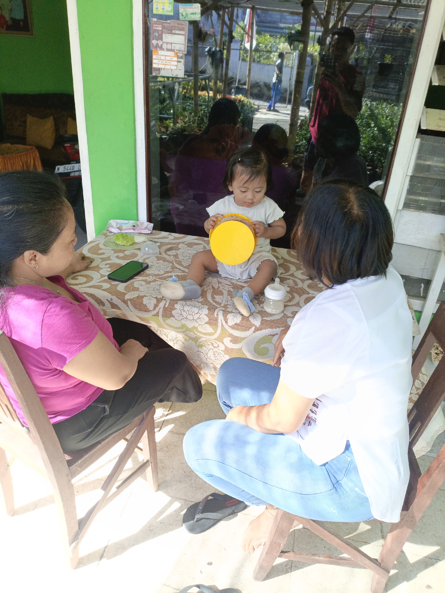 Two women and a toddler are seated around a table outdoors. The woman on the left wears a pink shirt and black pants. The woman on the right wears a white shirt and blue jeans. The toddler, wearing a white shirt and gray bottoms, sits on the table. The toddler holds a yellow object, possibly a toy. On the table are a pacifier, a baby bottle, and a green wipe. The table is covered with a patterned cloth. A cell phone rests on the table. The background shows a building with a glass window reflecting people and foliage. The scene appears to be in Batu, Indonesia, indicated by a license plate on a car partially visible on the left side of the image. The ground is paved with tiles, and sunlight casts shadows.