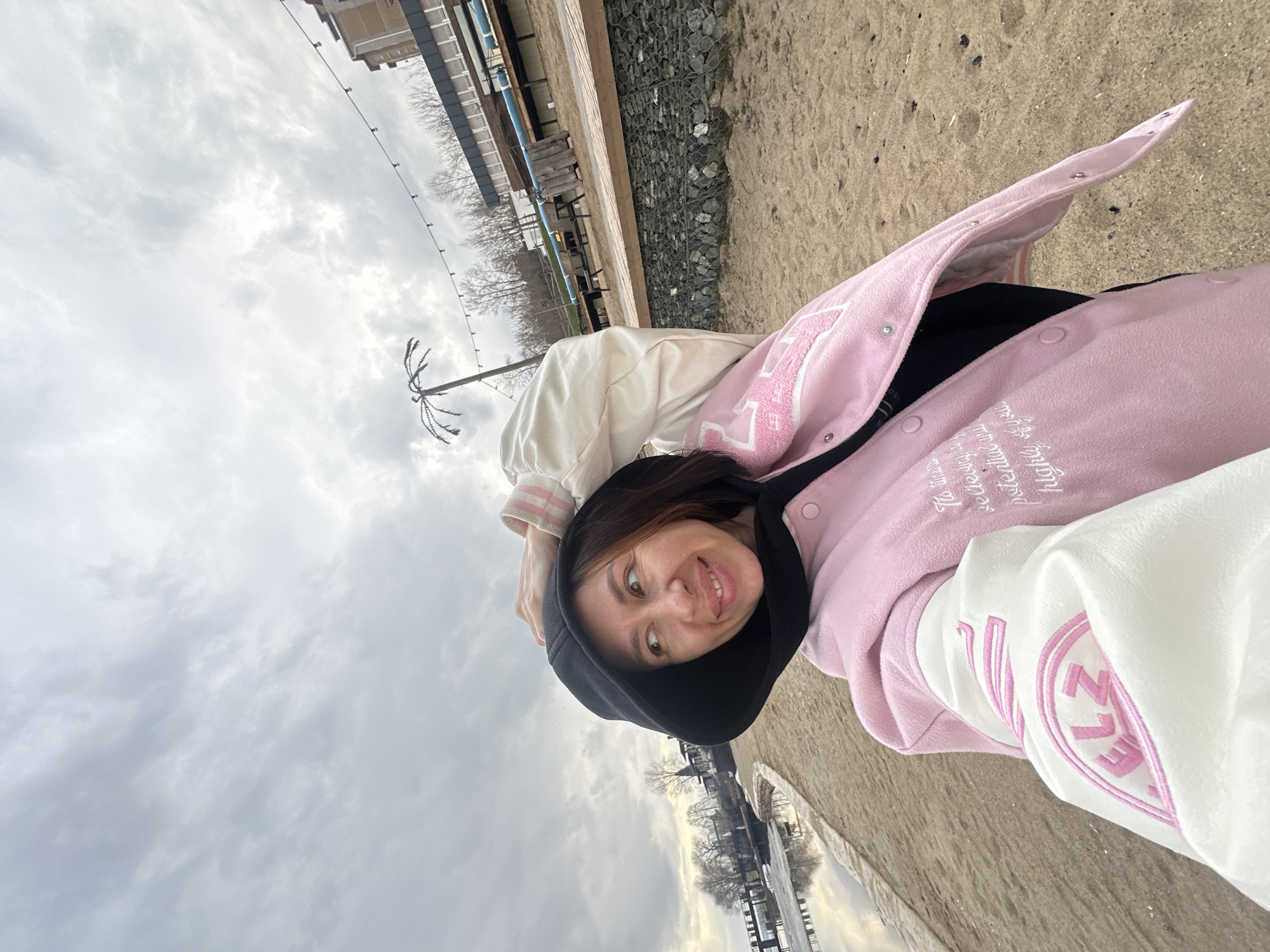 A woman is posing for a selfie at Votkinsk, Russia, near a body of water. She wears a black hooded sweatshirt underneath a pink and white varsity jacket, with the letter “LZ” embroidered on the sleeve. The background reveals a cloudy sky and bare trees lining the shore. In the distance, buildings and streetlights are visible. The ground appears to be sandy, with a gravel-covered area near a wooden structure.