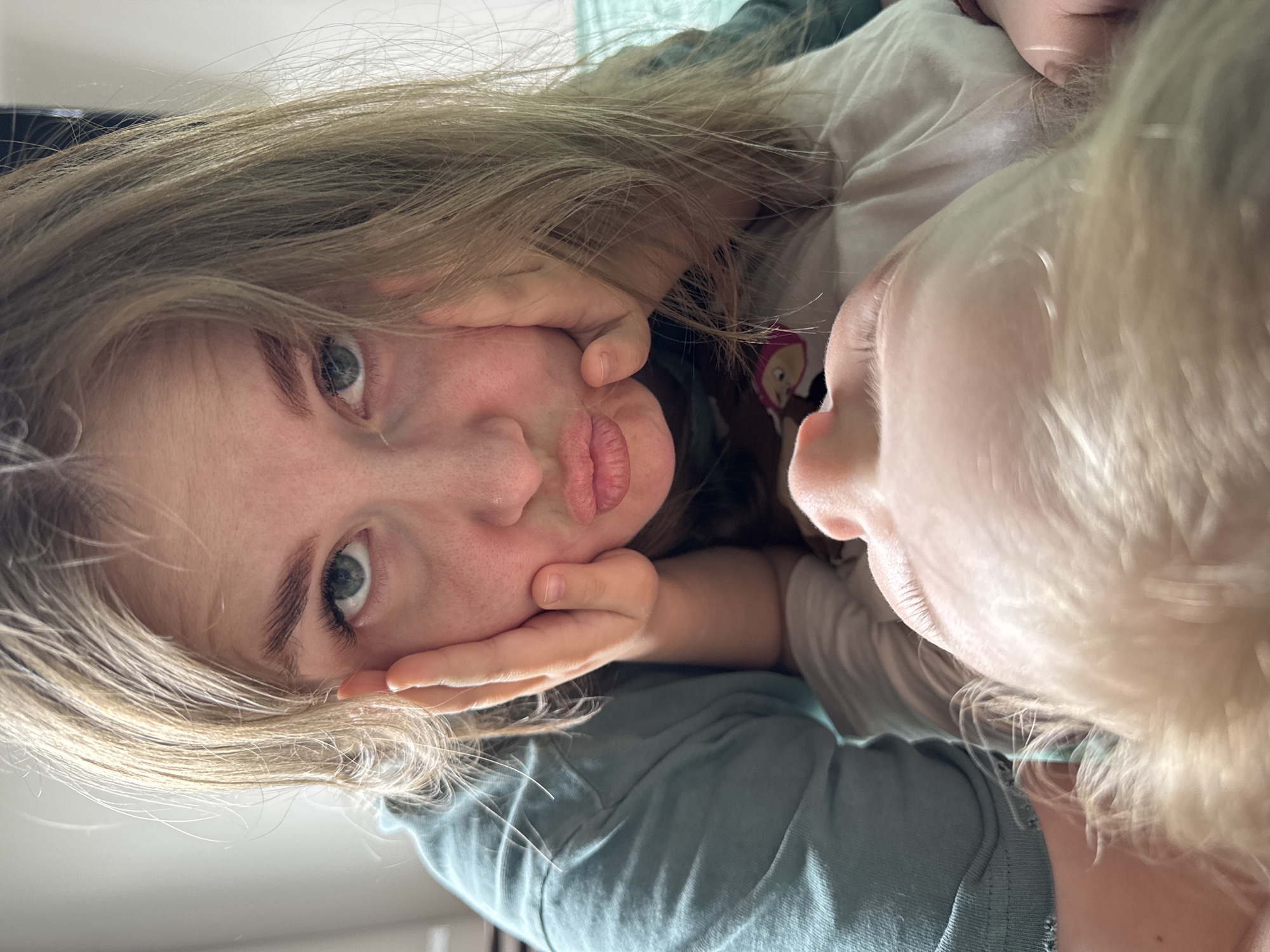 The image is a close-up, low-angle shot of two people. A young woman with light brown hair is on the left, looking at the camera with her hands positioned on her face, pulling her cheeks inward. Her lips are puckered in a kissing expression. The person on the right appears to be a child with fair skin and light blonde hair. Only part of their face is visible, including their nose and mouth. Both people appear to be lying down. The background is slightly blurred and indistinct, with hints of light-colored fabric and wall. There is no information to indicate this image was taken in the city of Ферма, Russia.