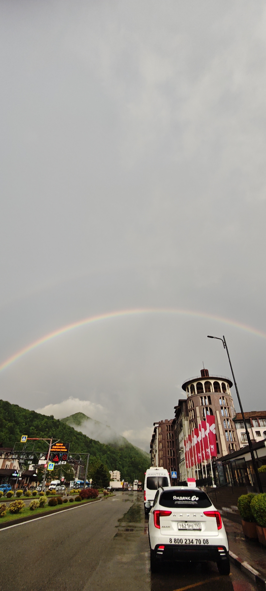 The image shows a street in Estosadok, Russia, with a prominent rainbow arching across the sky. The street is wet, reflecting the gray, overcast sky. In the foreground, a white car with "Яндекс Go" branding is parked on the right side of the street. Behind the car, a white van with "Mantera" written on it is also parked. On the right, a multi-story building with a distinctive round tower is visible. A row of flag-like banners in white and red adorns the building's facade. On the left, the scene transitions to a view of mountains partially obscured by clouds. A traffic signal is visible in the distance, near an intersection with pedestrian crossings. There are other vehicles in the distance, including cars and trucks. Along the street, there are landscaped areas with various plants and flowers. A street lamp is visible next to the building on the right.