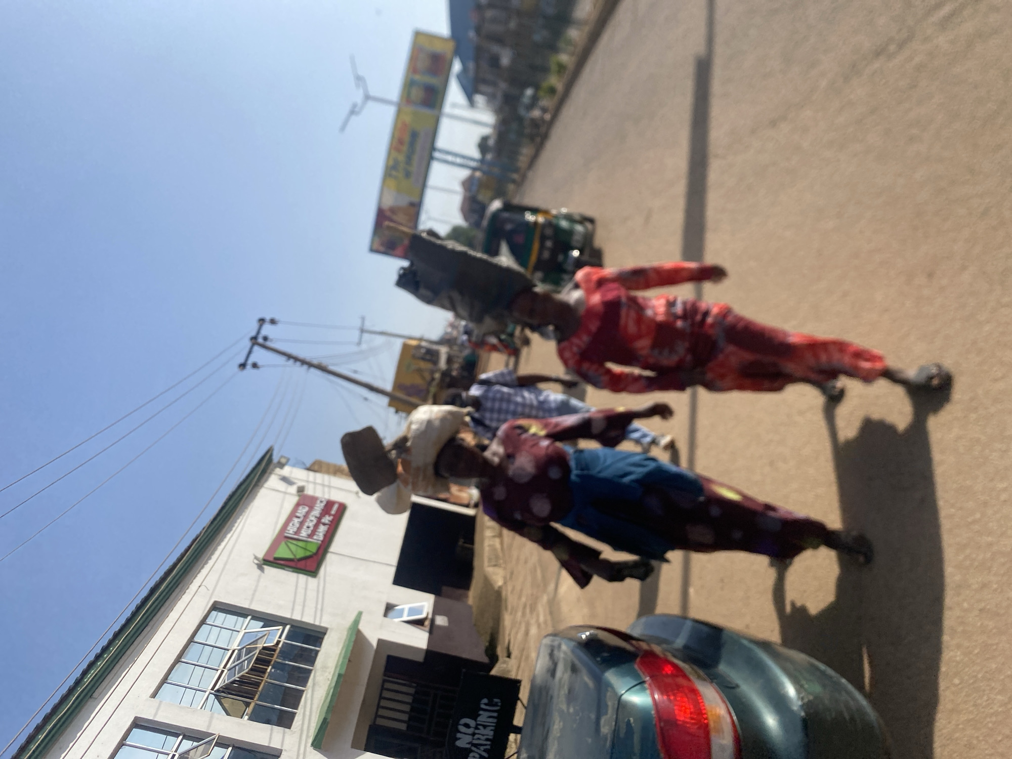 In Bukuru, Nigeria, two women are walking on a street, each carrying a large load on their heads. The woman on the left wears a dark red dress with white polka dots and a blue wrap around her waist. The woman on the right wears a red and white patterned dress. A dark green car is parked on the left side of the road, with a "NO PARKING" sign visible. A building with windows and a green trim is situated behind the car, bearing a sign that reads "HIGHLAND MICROFINANCE BANK PLC". In the background, there are more buildings, utility poles, and a bright blue sky. A three-wheeled auto-rickshaw is also visible on the street. The ground is a sandy, reddish-brown color.