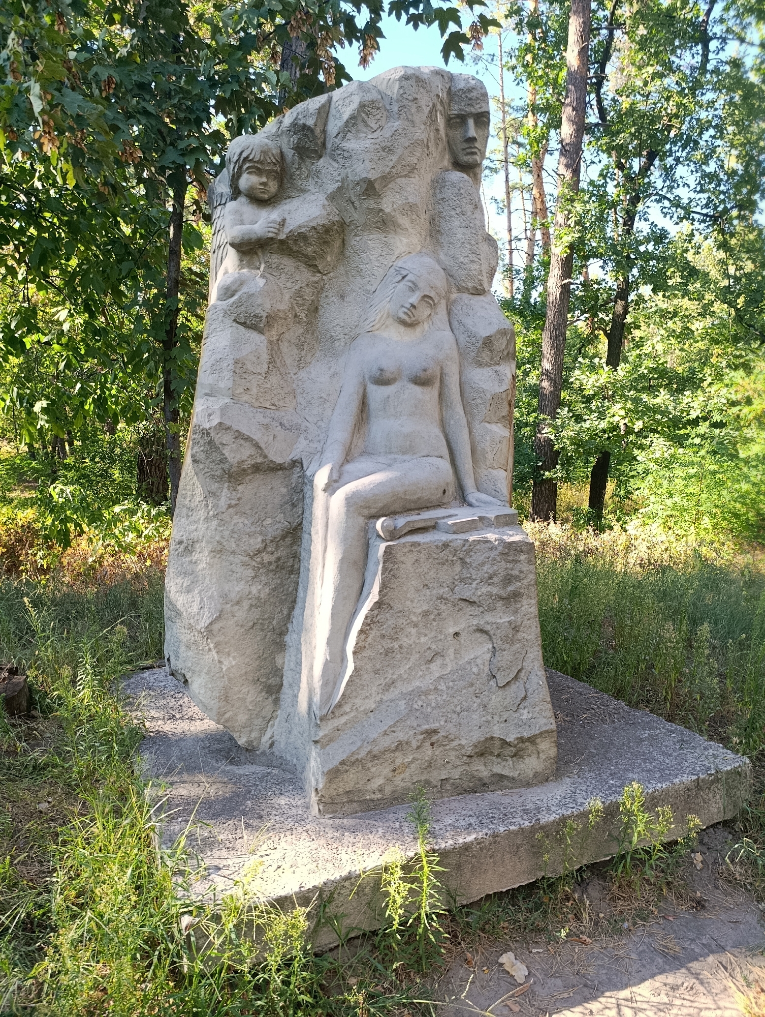 The image shows a large, light-colored stone sculpture set in an outdoor environment with trees and grass. The sculpture depicts three figures: a child in the upper left, a reclining female figure in the center, and a male face carved into the upper right of the stone. The child is looking downwards with arms crossed, while the female figure is posed with her head tilted back and her body partially exposed. The male face is looking to the side. The sculpture is positioned on a concrete base, and the surrounding area is covered in grass and some fallen leaves. The lighting suggests it is daytime with sunlight filtering through the trees. The overall scene appears to be in a park or wooded area.