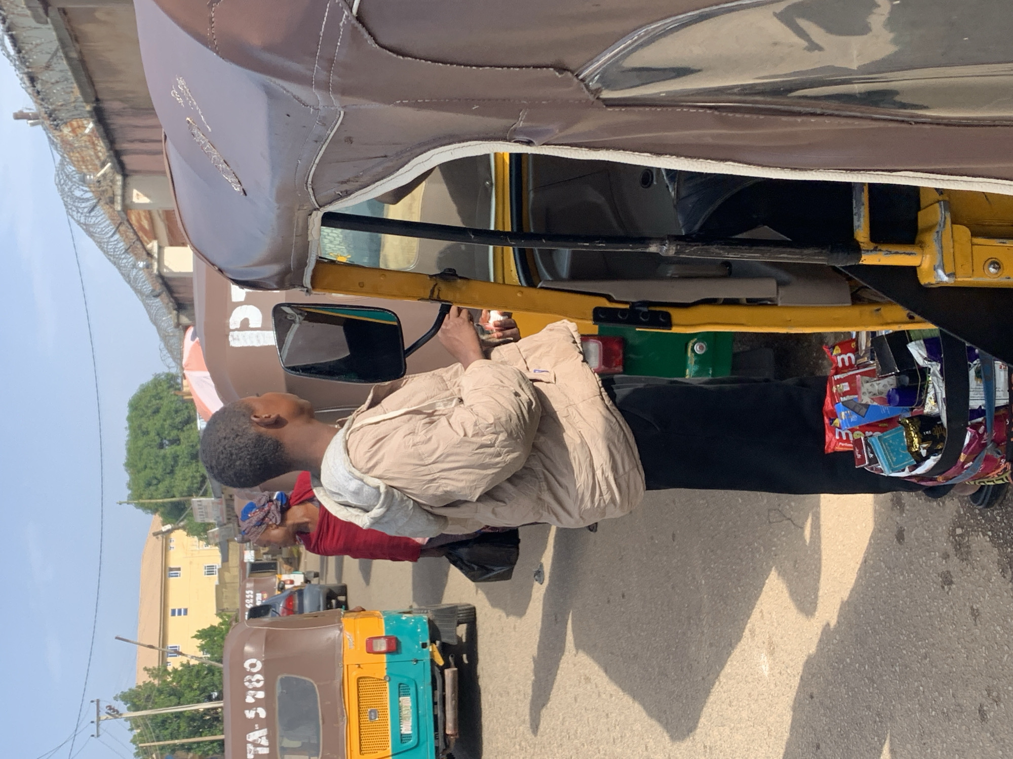 In Jos, Nigeria, a young person stands beside a brown and yellow auto-rickshaw, holding a small white object. They are wearing a light brown hooded jacket and black trousers. To the left of the auto-rickshaw, another auto-rickshaw with license plate TA-5780 is visible. Further in the background, a woman in a red headscarf and red garment stands with a black bag. The street is paved with asphalt, and shadows suggest a bright, sunny day. In the distance, a two-story building with a yellow facade and white window frames stands under a clear sky. A wall topped with barbed wire runs along the right side of the image. A basket filled with various packaged snacks rests at the feet of the young person in the foreground.