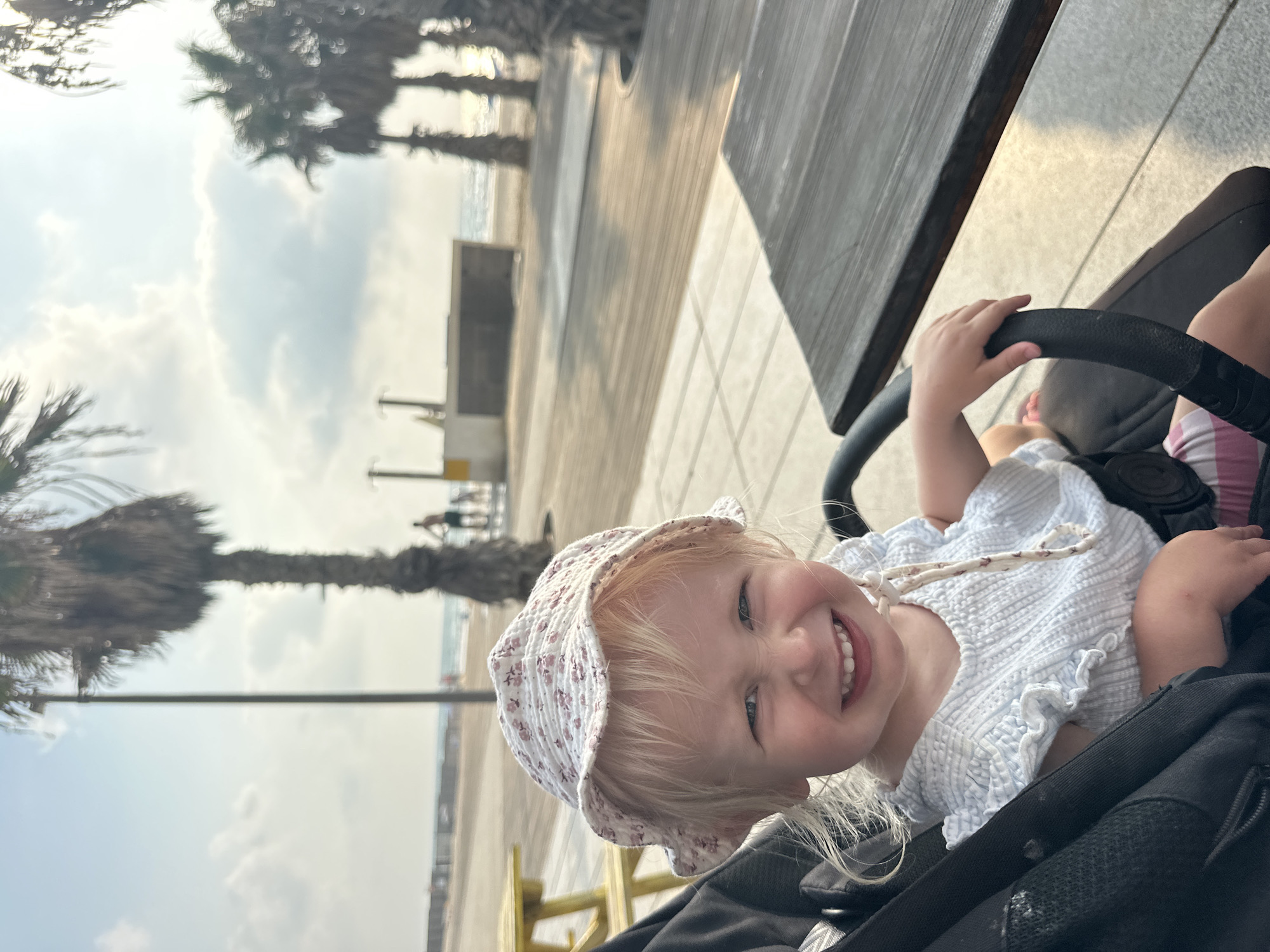 This image captures a close-up of a young child, likely a toddler, sitting in a stroller. The child has blonde hair and is wearing a light-colored sun hat with a floral pattern and a white, textured blouse. They are smiling broadly, with their hand resting on the stroller's handlebar.
The background suggests an outdoor setting, possibly a boardwalk or promenade next to a beach. Palm trees are visible, and the sky is cloudy, indicating it might be daytime with mixed weather. In the distance, a body of water, likely the sea, can be seen, along with a sandy beach. There are some structures and people faintly visible in the background, hinting at a public space. The ground appears to be paved with light-colored tiles. A wooden table or bench is also present in the upper right portion of the frame.
The overall mood is cheerful, primarily due to the child's joyful expression. The lighting suggests it's likely daytime, possibly late afternoon given the quality of light. The presence of palm trees and the sea suggests a coastal location, and the provided context specifies Bat Yam, Israel. No visible text is present in the image.