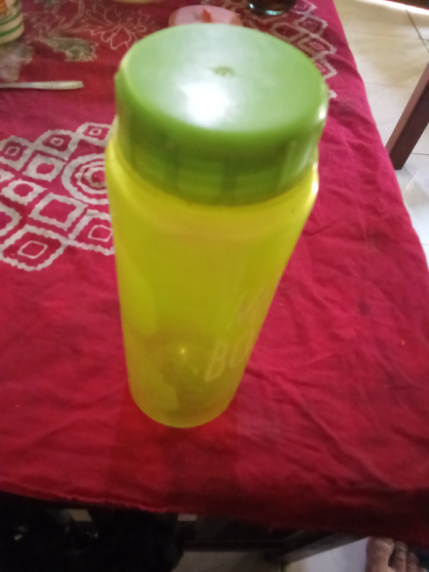 A bright yellow plastic water bottle with a green lid stands upright on a red textured tablecloth. The tablecloth has a white patterned design. The background is blurred, but it appears to be an indoor setting with wooden furniture. The image quality is poor, with a low resolution and lack of sharp focus, making it difficult to discern finer details. The scene is static and there are no discernible actions or interactions taking place. The water bottle appears to be the main subject of the photograph. The location in the city of Pemalang, Indonesia, is not visually evident from the image itself.