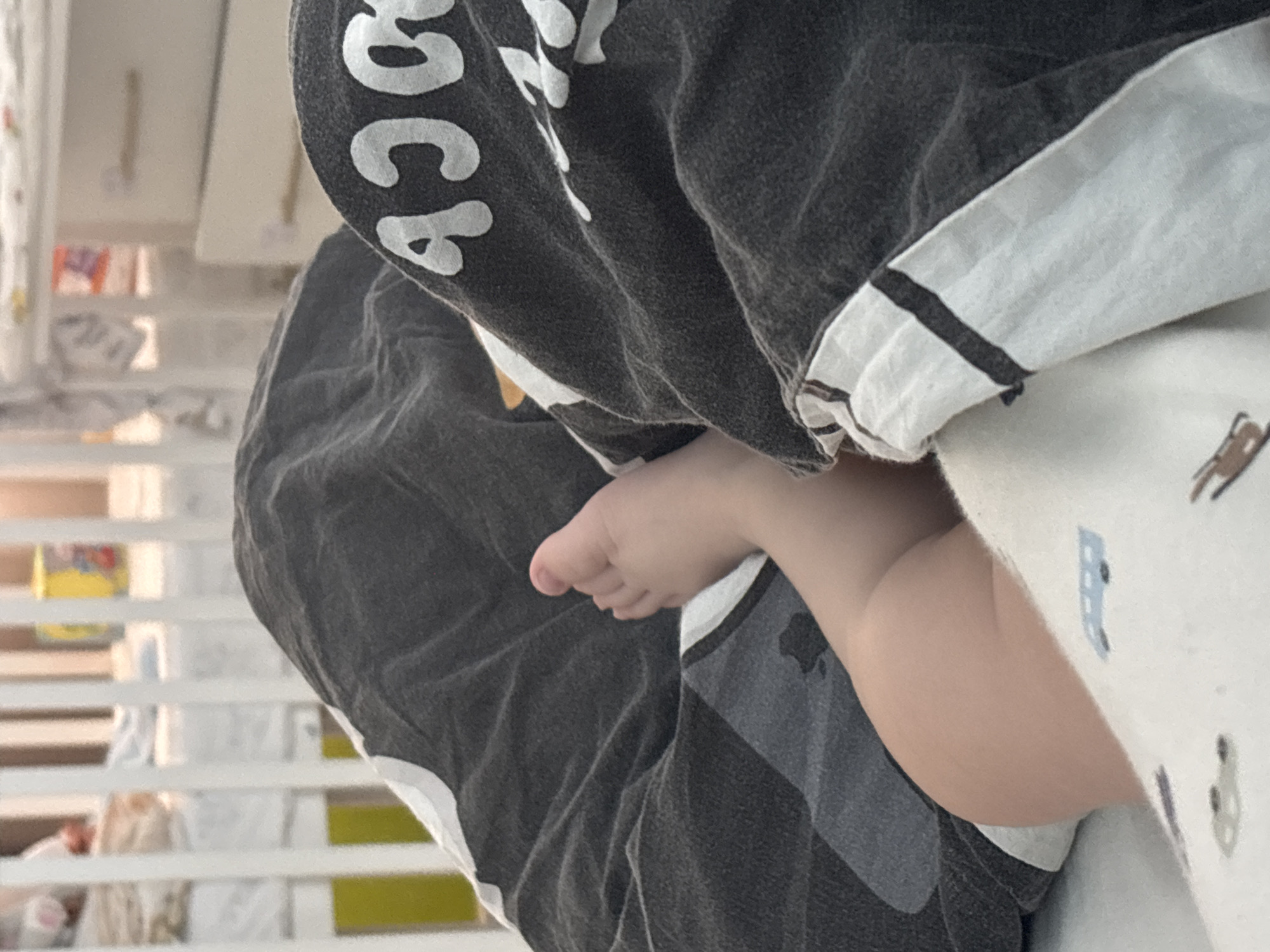 A close-up shot captures a baby's foot peeking out from under a patterned blanket. The blanket is partially obscured by a dark grey t-shirt. The background features a white crib with vertical slats. The image was likely taken indoors. There is no information to suggest the image location is Раздолье, Russia.