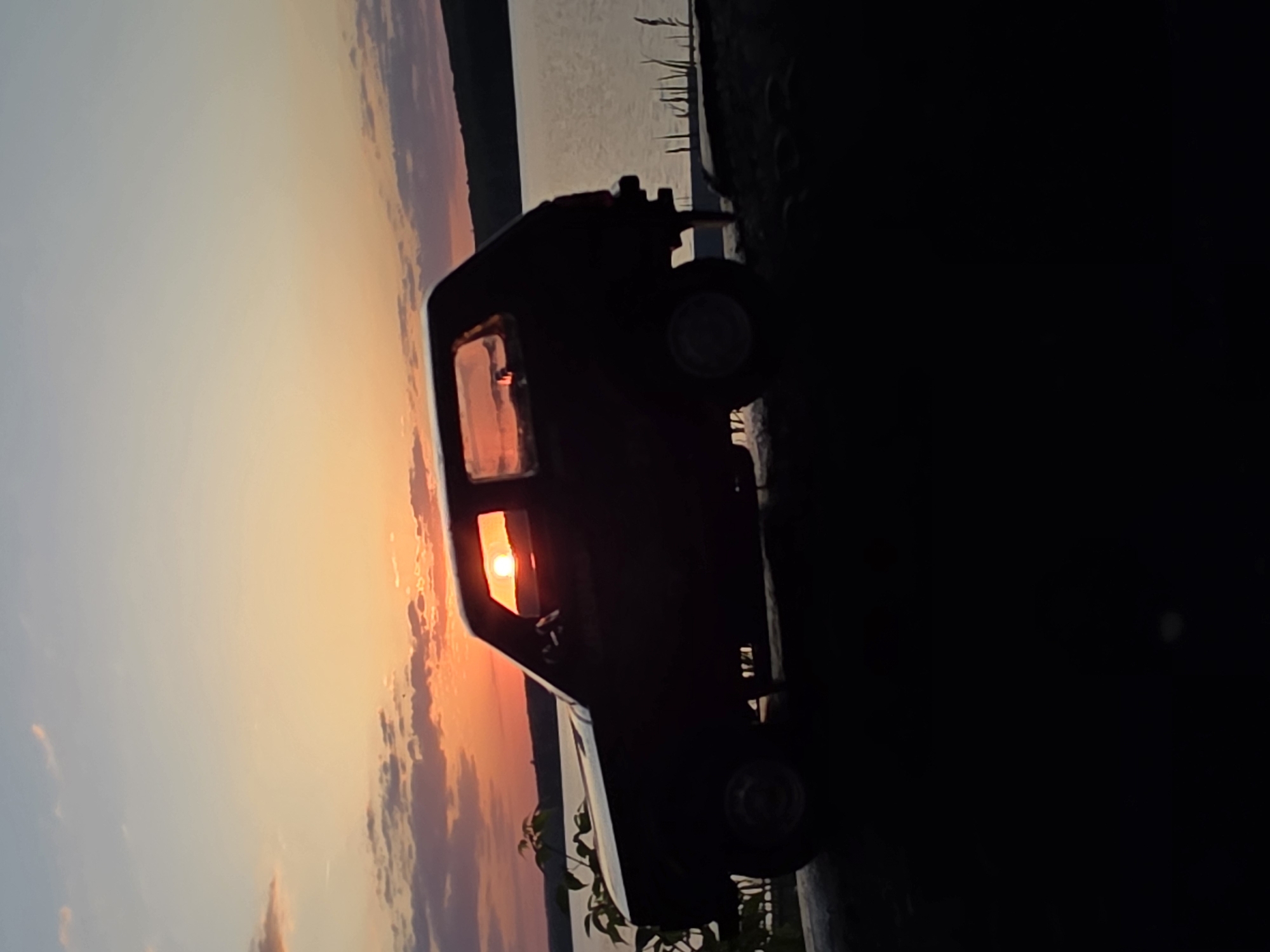 The image shows a dark silhouette of a vehicle, possibly a compact car or small truck, parked near what appears to be a shoreline or waterfront. The sky in the background features a vibrant sunset, with colors ranging from orange and pink to pale blue. The setting is likely near a body of water, given the smooth expanse visible in the background, possibly the Kama River, typical of Chaikovsky, Russia. Some vegetation is visible at the bottom of the frame, suggesting a grassy or natural area near the road. The image is slightly rotated, tilting the vehicle and the horizon. It's difficult to determine the exact location within Chaikovsky without additional context or landmarks.