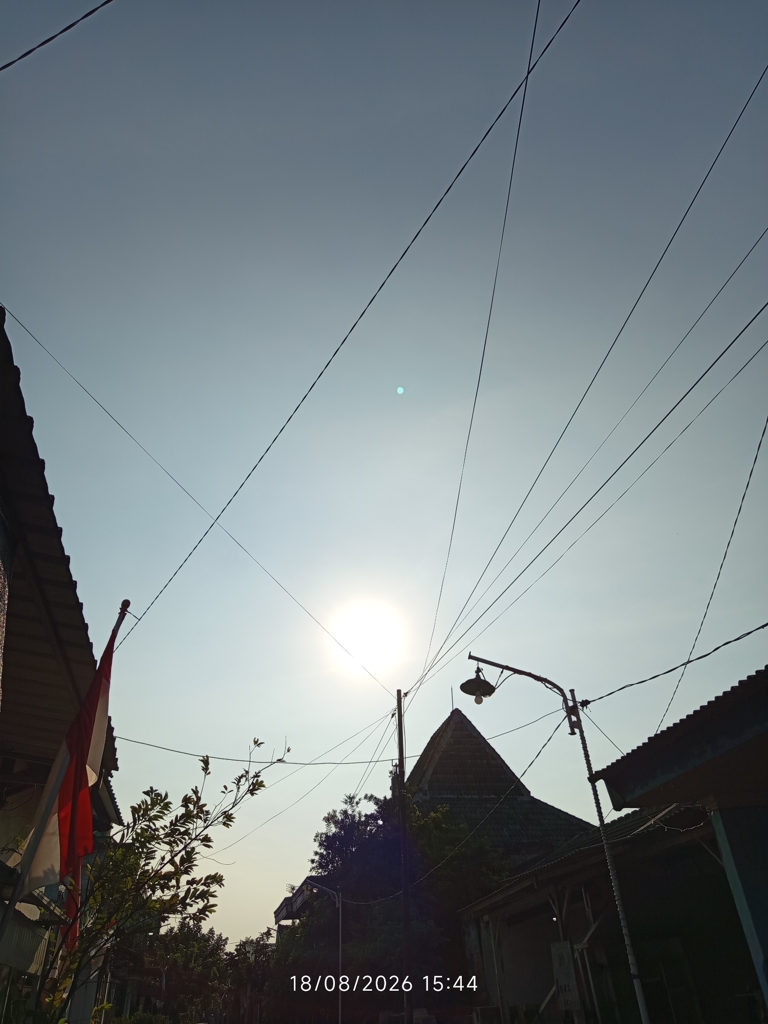 An outdoor, low-angle shot captures a residential street in Canggu, Indonesia. The sky is a pale blue, dominated by a bright, hazy sun positioned slightly left of center. A complex network of utility wires crisscrosses the frame, some appearing to converge near a street lamp with a visible bulb. On the left side, a red and white Indonesian flag is partially visible, attached to a pole near the edge of a tiled roof. To the right, another building with a corrugated metal roof is visible. Trees and foliage are interspersed between the structures. The lower right corner of the image displays a timestamp: "18/08/2026 15:44". No people are visible in the scene.