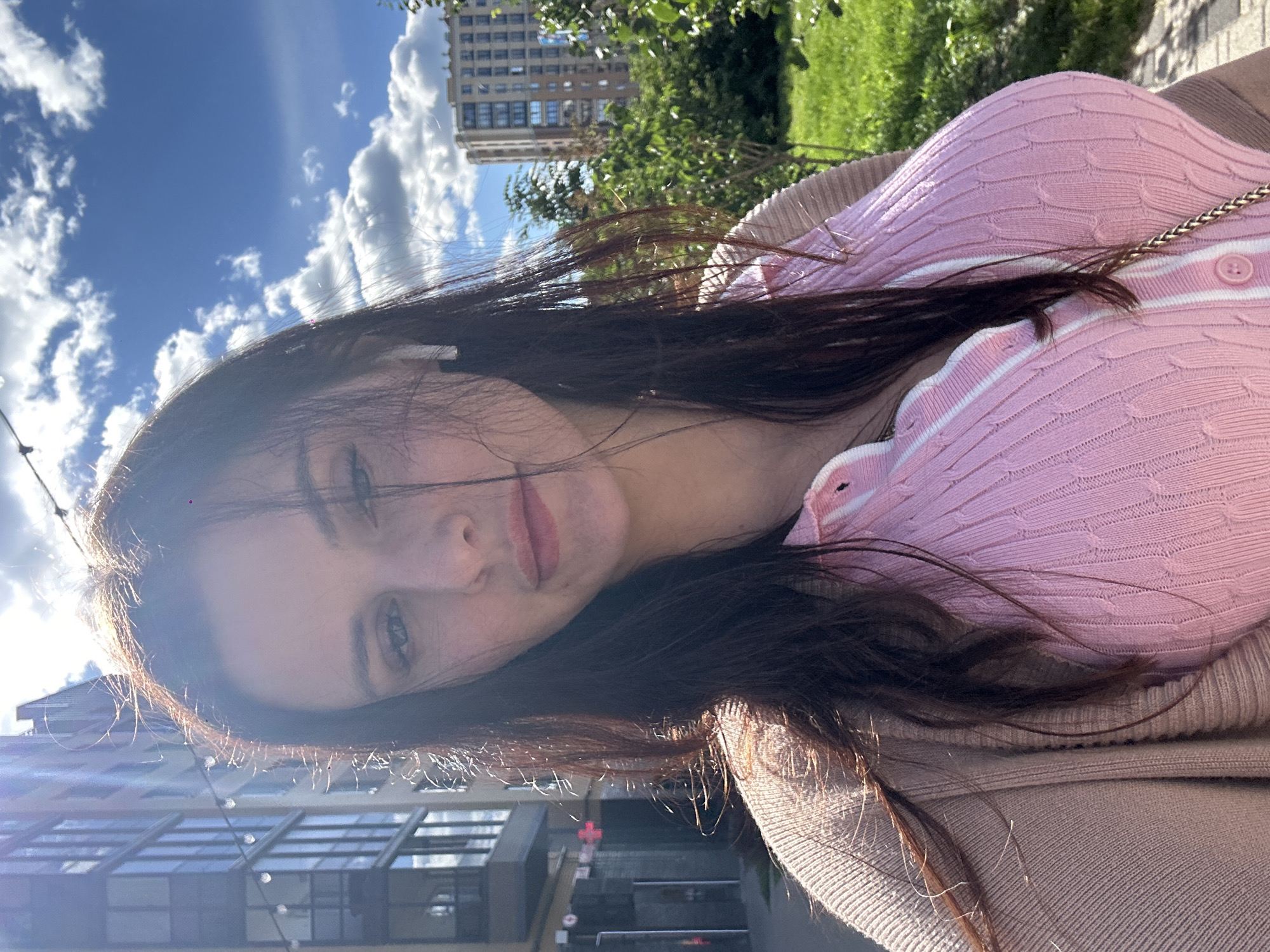 This media file is a selfie of a young woman taken outdoors. She has long, dark brown hair that frames her face and is partially obscuring her features. She is wearing a pink knitted cardigan with white trim and a gold chain visible over her shoulder. Her expression appears relaxed, with a slight smile.
The background is blurred but suggests an urban setting with buildings and greenery. The lighting indicates it is daytime, with bright sunlight casting shadows and creating lens flare, particularly in the lower left portion of the image. The sky is visible with some scattered white clouds. The overall impression is casual and bright, likely taken on a pleasant day. There is no visible text in the image.