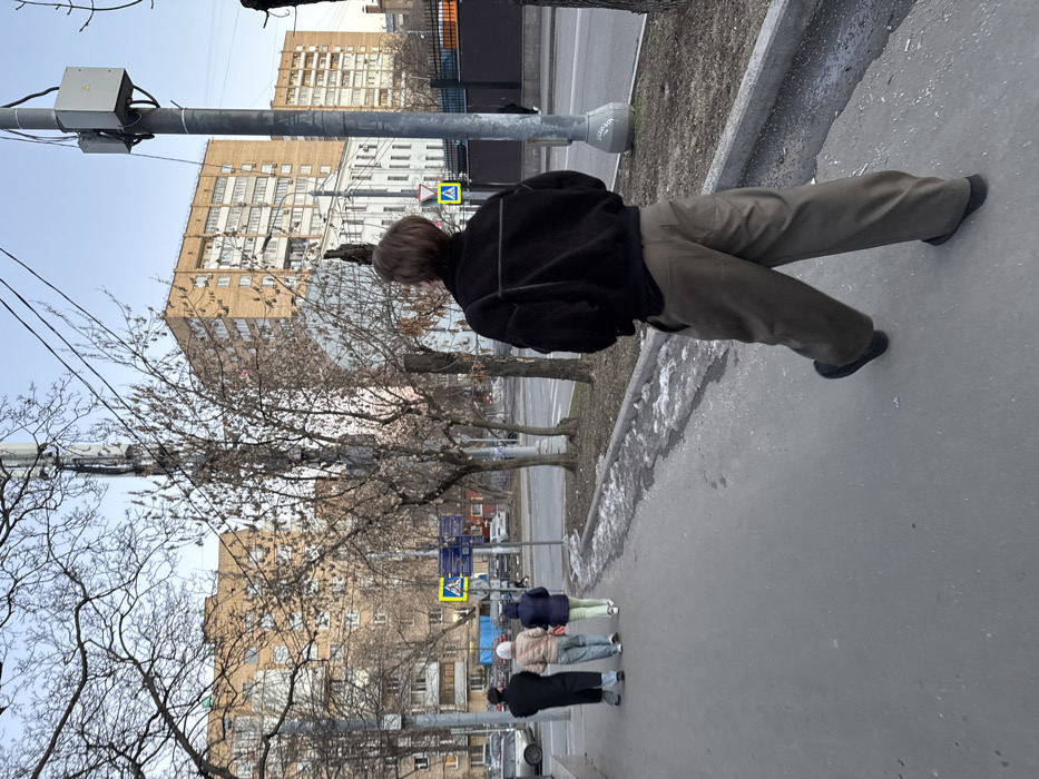 This image depicts a late afternoon or early evening street scene in Moscow, Russia, during late winter or early spring, characterized by bare deciduous trees and residual dirty snow/ice along the roadside. The sky is clear and light blue.
In the immediate foreground, facing away from the camera and moving towards the right, is a person with short light brown hair. They are wearing a dark, possibly black, bomber-style jacket with horizontal seam details across the back, and loose-fitting, wide-leg olive-green trousers paired with dark shoes.
Further along the concrete sidewalk, three additional individuals are walking away from the camera. The leftmost person wears a dark, likely black, long coat and a dark head covering. The middle person is dressed in a light-colored jacket and light trousers or a skirt. The rightmost person wears a dark blue hooded jacket and light green trousers.
The midground features the multi-lane paved road, partially obscured by trees. Utility poles with attached electrical boxes and a tall pole equipped with multiple cellular antenna arrays are visible. Patches of melting snow and ice lie along the curb.
In the background, a row of buildings lines the street. On the far left, a multi-story tan brick residential building with balconies is prominent. To the center-right, a lighter-colored, possibly white or pale yellow, multi-story building with a darker roof and distinctive window shapes (some arched) stands between the brick residential structures. Another similar tan brick residential building is visible on the far right. A yellow triangular pedestrian crossing sign with a black border and a black walking person symbol is visible above blue street signs, indicating a pedestrian crossing. Multiple vehicles are visible on the street.