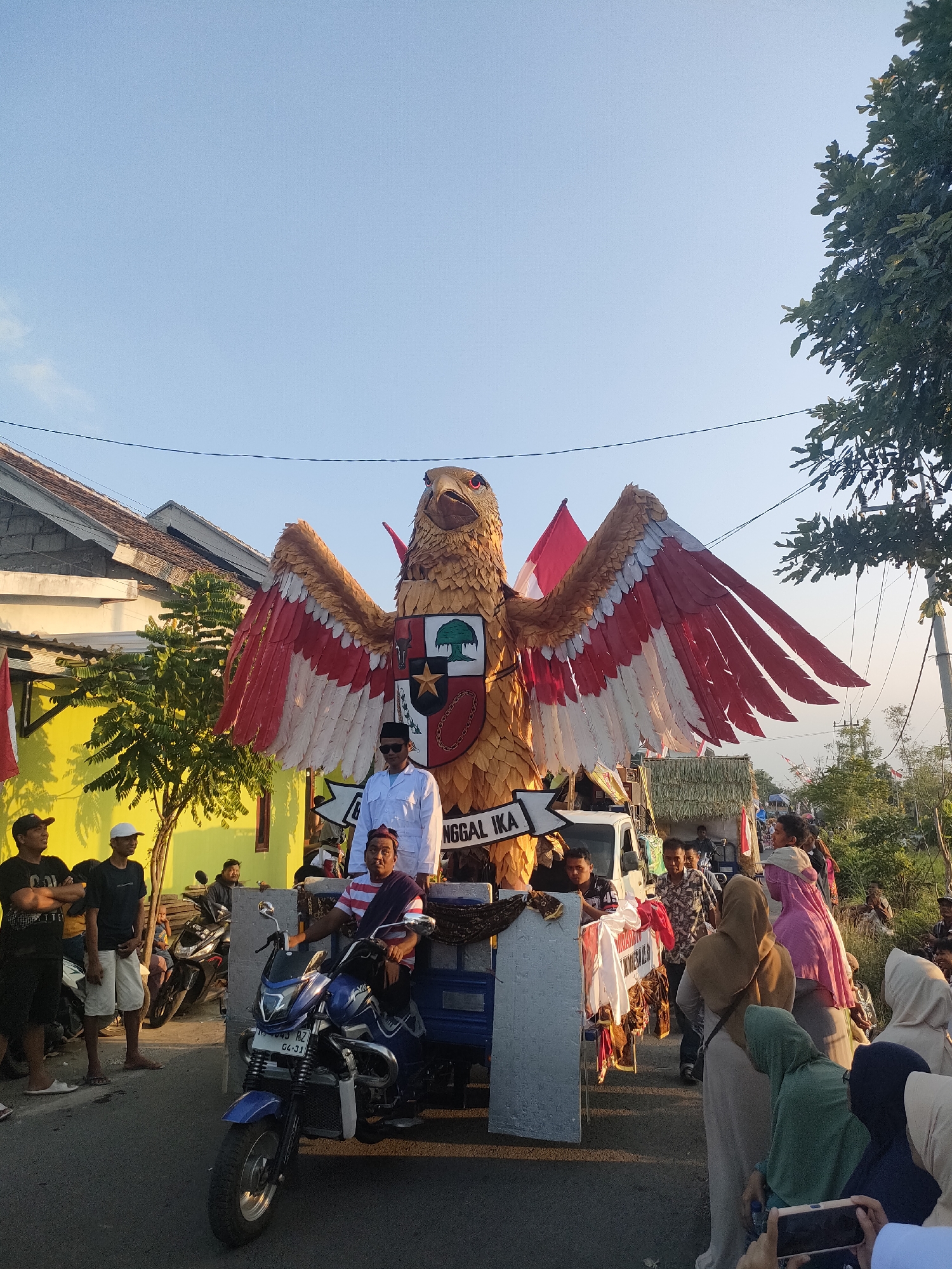 In Tongaswetan, Indonesia, a large, elaborate eagle float, adorned with red and white wings, is prominently displayed on a trailer. The eagle's chest bears a shield with the Indonesian national symbol, Garuda Pancasila. Several individuals are positioned on the float, including a man in a white shirt and sunglasses, and another man wearing a striped shirt and a red head covering. In front of the float, a blue motorcycle-drawn cart is visible, with a man seated on it. A crowd of spectators, many wearing headscarves, observes the spectacle from the roadside, which features houses and trees. The scene appears to be a parade or a public celebration, with the clear blue sky indicating daytime.