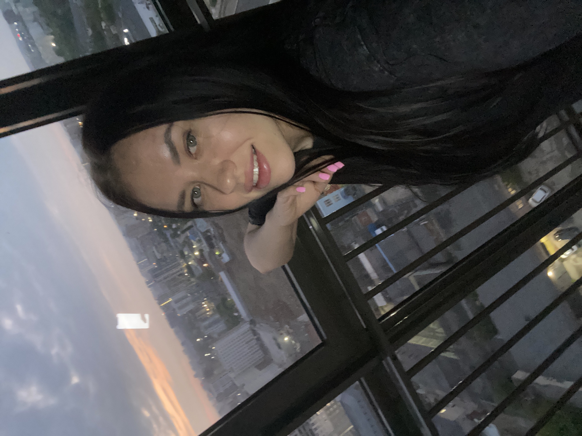 A medium shot, taken from an elevated position, shows a young woman with long, dark hair and green eyes. She is looking towards the camera and smiling, with her right arm resting on a metal railing. The background reveals a city skyline at dusk, with buildings and lights visible. The sky is a mix of grey clouds and a soft orange glow from the setting sun. A rectangular white object, possibly a reflection or an advertisement, is visible in the sky. The overall scene suggests an urban environment in Perm, Russia.