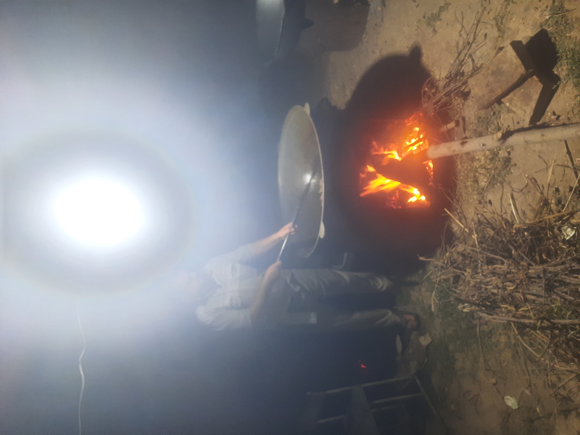 In the city of Vosobad, Tajikistan, a person is seen standing next to a large, dark pot, possibly cooking. The scene is illuminated by a bright, overhead light source that creates a halo effect and obscures details. A fire burns beneath the pot, casting a warm glow on the surrounding dry twigs and branches. The ground appears to be dirt or sand. A chair and another pot are visible in the background. The overall atmosphere is dimly lit and hazy due to the intense light and smoke.