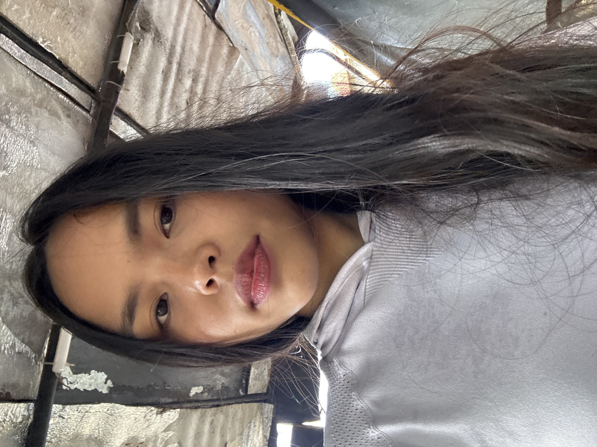 A selfie is taken of a young woman with long, dark hair and light skin. She is wearing a light gray hooded sweatshirt. Her lips are painted with a reddish lipstick. She is looking directly at the camera with a neutral expression. The background appears to be indoors, with a corrugated metal ceiling and some wires visible. The lighting suggests it may be daytime. The location is described as being in the city of Davao City, Philippines.