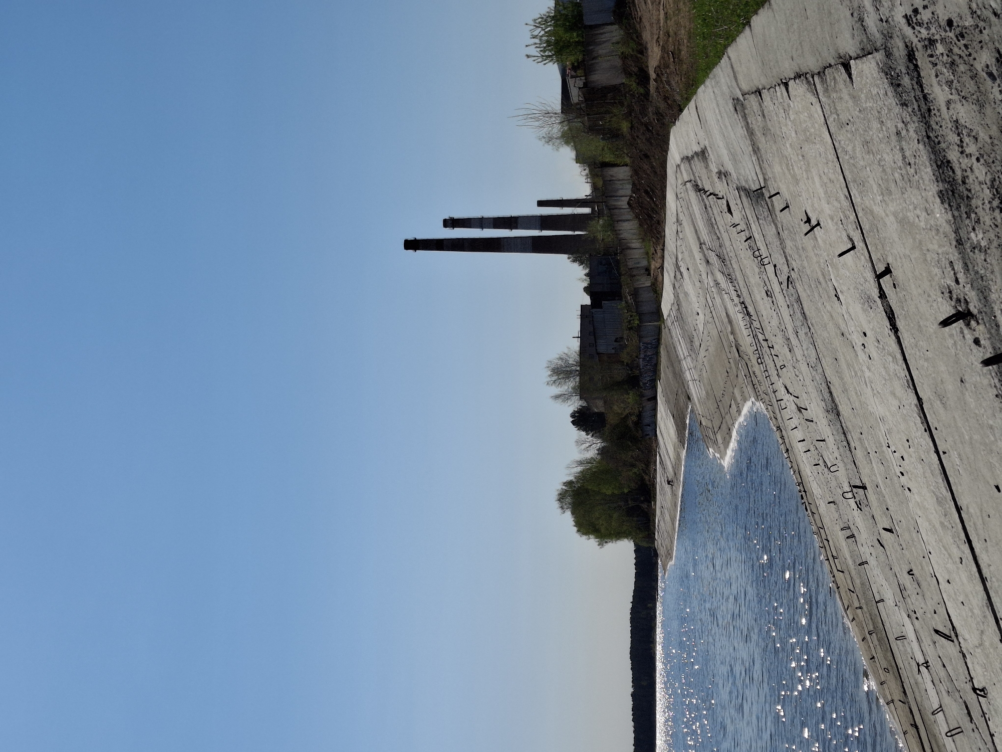 The image shows a view from the embankment in Chaikovsky, Russia. The concrete embankment edges the water, with the reflections of sunlight creating a shimmering effect on the surface. Beyond the embankment, there is vegetation and old industrial buildings, including brick smokestacks, typical of industrial zones. The sky is clear and blue. The photograph is taken at an angle, which makes the concrete embankment appear nearly vertical in the frame.