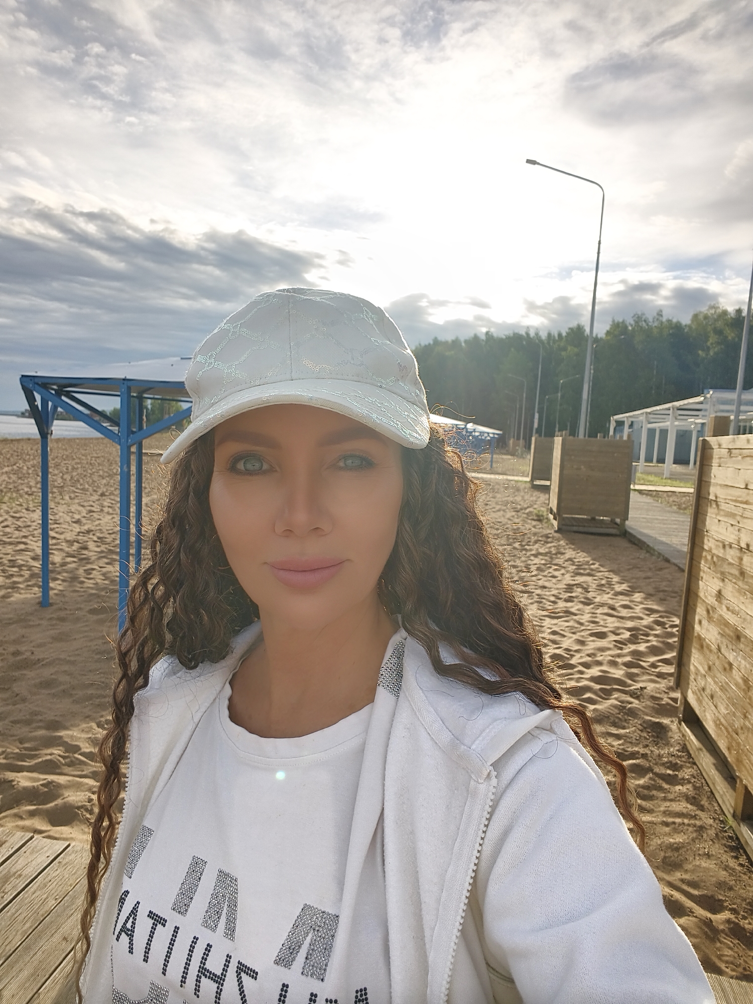 A woman with long, curly brown hair and striking blue eyes is taking a selfie outdoors on a sandy beach. She is wearing a white baseball cap and a white hooded jacket over a white t-shirt. The t-shirt has dark text with a sparkly embellishment. She has light pink lipstick on and is smiling softly.
The background shows a beach with sand dunes and a wooden walkway. There are some beach structures, including a blue shelter with a roof and poles, and wooden changing cubicles. In the distance, there are trees and a body of water. Several streetlights are visible, suggesting a developed area. The sky is partly cloudy with the sun shining through, indicating it is daytime, likely late afternoon or early evening due to the warm lighting.
The overall mood is relaxed and pleasant, with the woman appearing to enjoy a casual outing by the water. The location context is Chaikovsky, Russia. There is no discernible activity other than the selfie being taken.