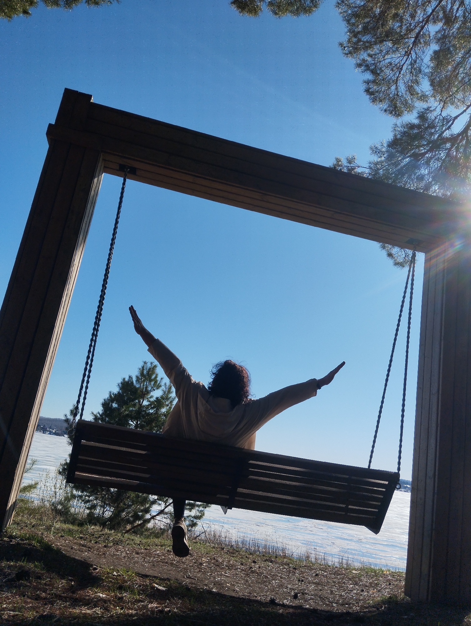 A woman sits on a swing in Votkinsk, Russia, with her arms outstretched. The swing is made of dark wood and hangs from a wooden frame. The chains connecting the seat to the frame are black. The woman is wearing a light-colored jacket. Behind the swing is a view of what appears to be a frozen body of water covered in snow. The sky is clear and blue. Trees are visible around the swing, with the sun casting a lens flare in the upper right corner of the image. The ground around the swing is covered in grass and dirt.