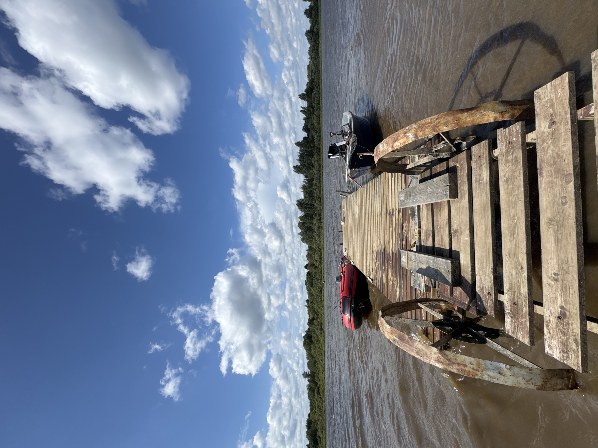A wooden pier extends into a body of water under a blue sky with scattered white clouds. Two boats are moored to the pier: a red inflatable boat with fishing rods visible and a blue motorboat with a black seat. The water appears murky brown, and a line of green trees forms the distant shoreline. The pier is constructed of weathered wooden planks and features rusty metal supports with wheel-like structures on either side. The scene is set on a sunny day.