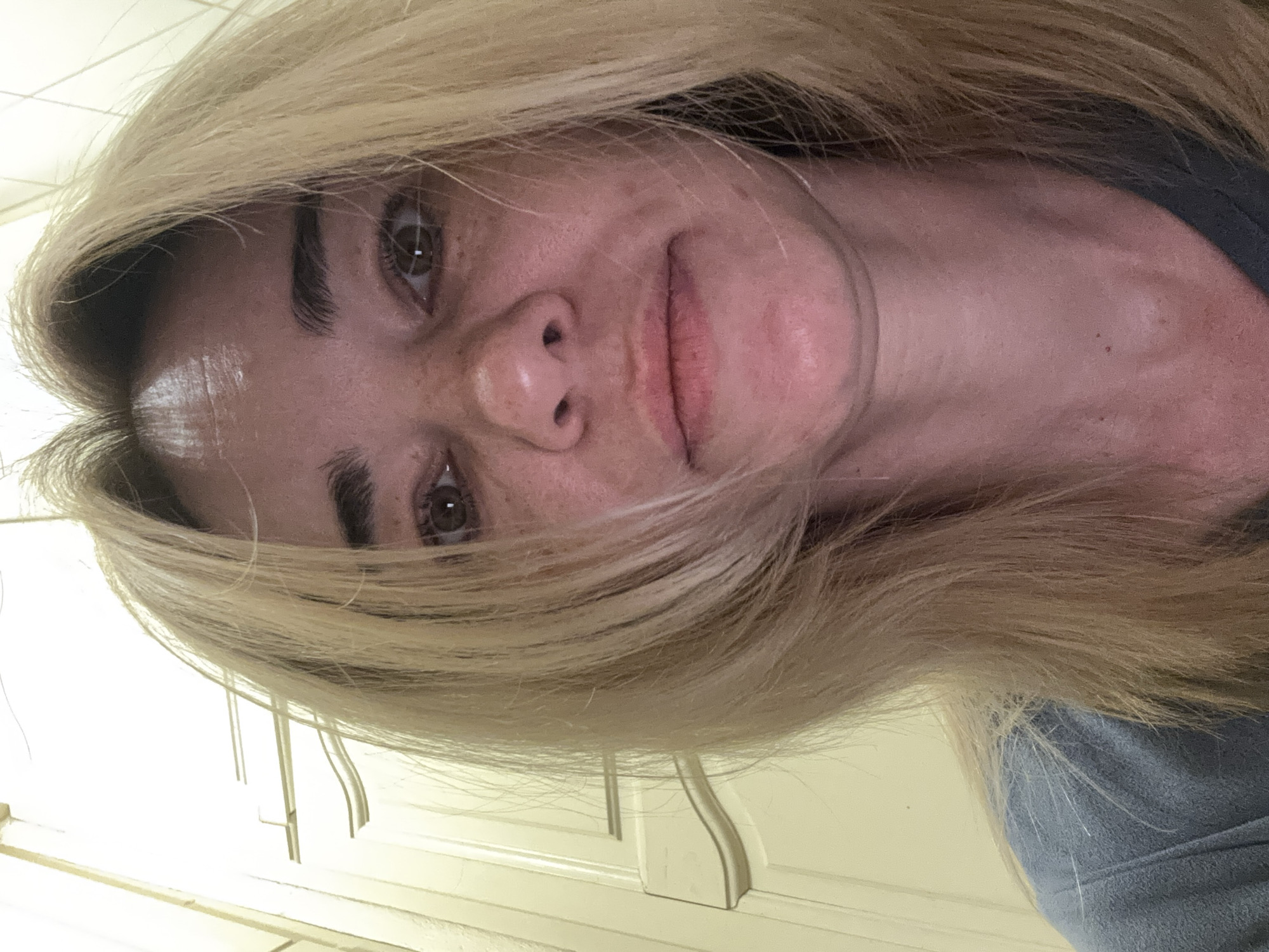 A close-up, rotated selfie of a woman with light blonde hair is presented. Her head is tilted, framing her face with strands of hair that partially obscure her forehead. Her eyes are brown. She has fair skin with freckles and a neutral expression. The background consists of white or light-colored paneled cabinets. The location is Unknown, Unknown.