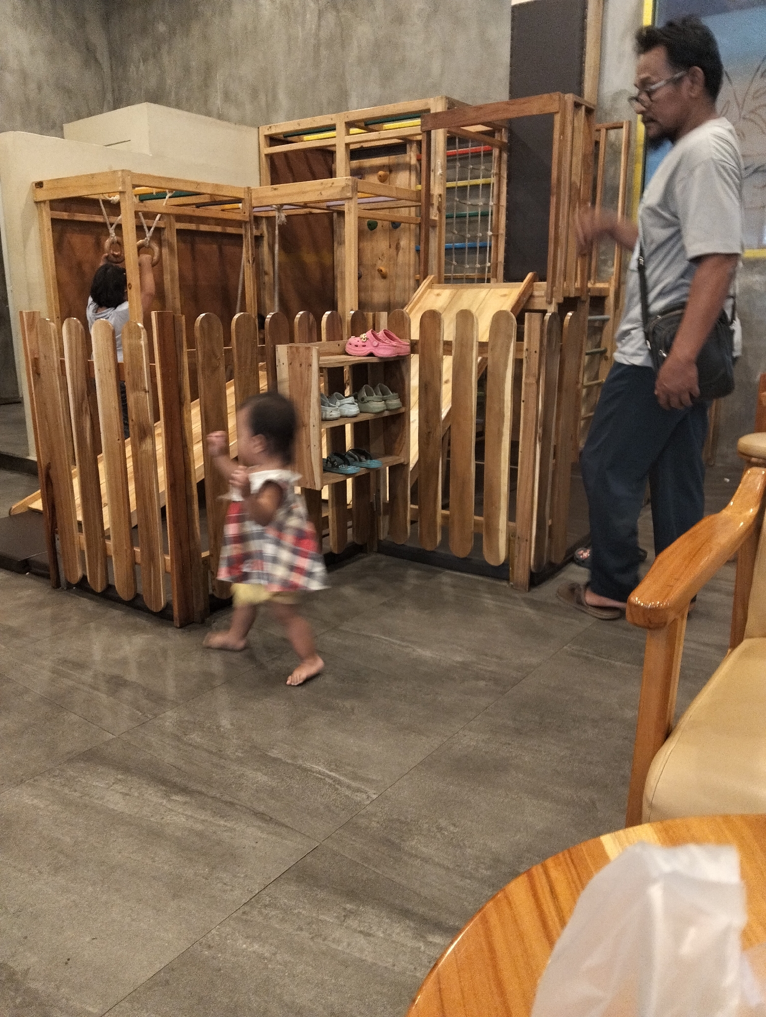 The image depicts an indoor play area with a wooden climbing structure and fence. A young child, wearing a plaid dress and yellow shorts, is walking barefoot on a tiled floor. An adult male, wearing a grey t-shirt, dark pants, and glasses, stands in the background. Several pairs of shoes are visible on shelves within the play structure. The overall environment suggests a children's entertainment facility. There is no information in the image to place the location in Sumberberas, Indonesia.