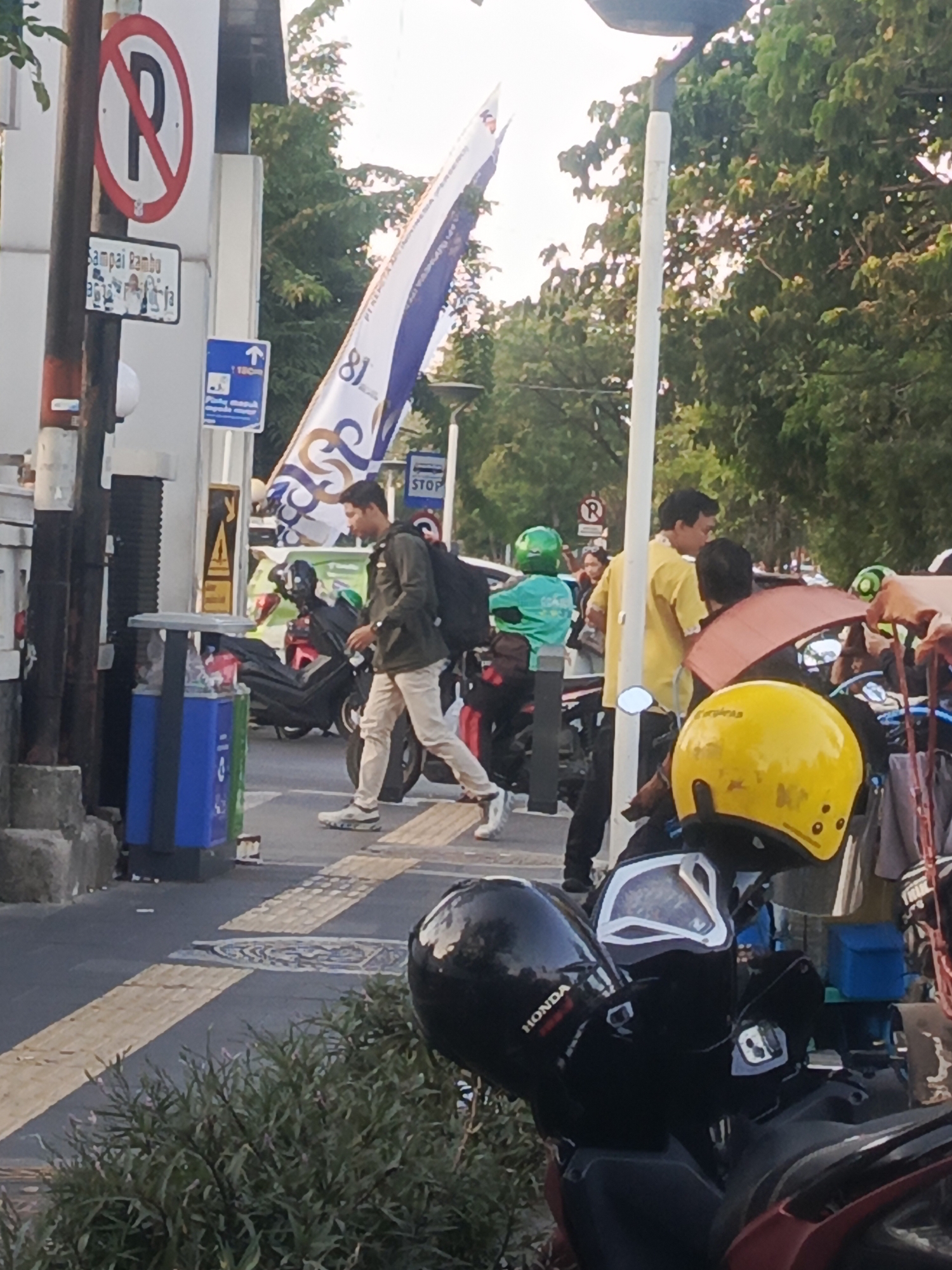 The image depicts a street scene in Surabaya, Indonesia, likely during daylight hours. A man wearing a green jacket and beige pants is walking across a pedestrian crossing. He carries a backpack and a drink in his hand. Several motorcycles are parked or waiting, with some riders wearing helmets, including a prominent yellow helmet in the foreground. A banner with the number "81" and blue and white motifs is visible, likely related to an event or celebration. Other signs include a "no parking" sign and a directional sign with an arrow and "100m". A trash receptacle is located on the left side of the image. Trees line the background, and the sky suggests a bright, possibly hazy, atmosphere. The presence of Indonesian signage confirms the location.