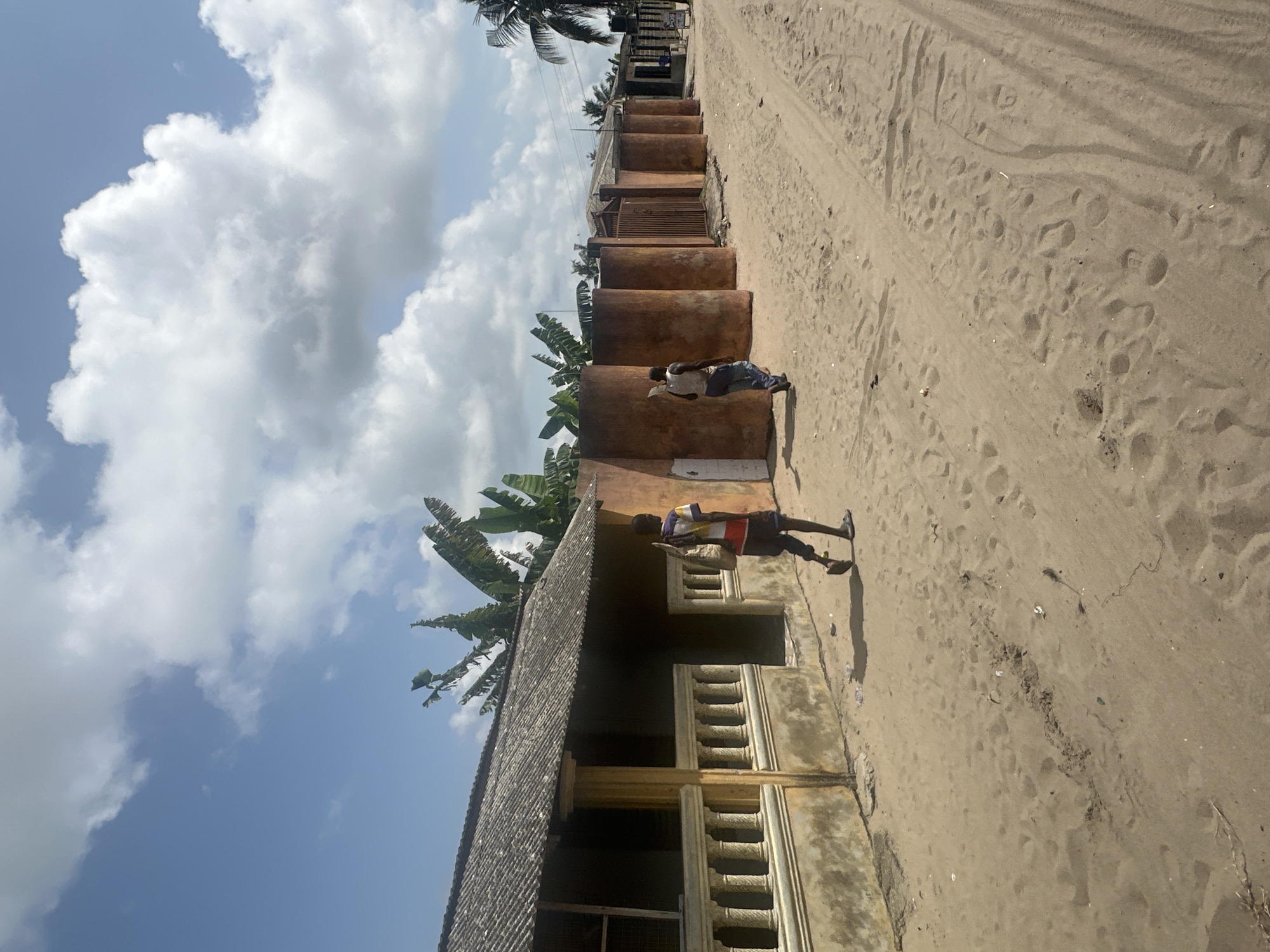 The image captures a sunny outdoor scene along a sandy or dirt road, typical of a rural or semi-urban setting, consistent with Badagry, Nigeria. The perspective is looking down a street or pathway, with buildings lining one side and a wide, sandy track on the other.
In the upper left, a bright blue sky is adorned with large, fluffy white cumulus clouds, indicating clear weather. The sun appears high, casting strong, distinct shadows, suggesting it's midday or early afternoon.
Along the left side of the frame, a row of weathered, single-story buildings extends into the distance. The building closest to the viewer features a porch or veranda with a balustrade and pillars, topped by a corrugated metal roof. Further down, several large, cylindrical, reddish-brown structures, possibly water tanks or large decorative pots, are arranged in a line. Lush tropical vegetation, including prominent banana plants and palm trees, grows around and behind these structures and buildings.
Two individuals are visible. Closer to the foreground building, a younger male wearing a multi-colored striped t-shirt and dark shorts stands, facing away from the camera. Further down the path, another person, an adult male, leans against one of the cylindrical structures. He is dressed in a white short-sleeved shirt and blue trousers and appears to be looking down intently, possibly at a book or phone.
The sandy ground on the right shows numerous footprints and tire tracks, suggesting active passage. No discernible text is visible on any signs or objects within the image.