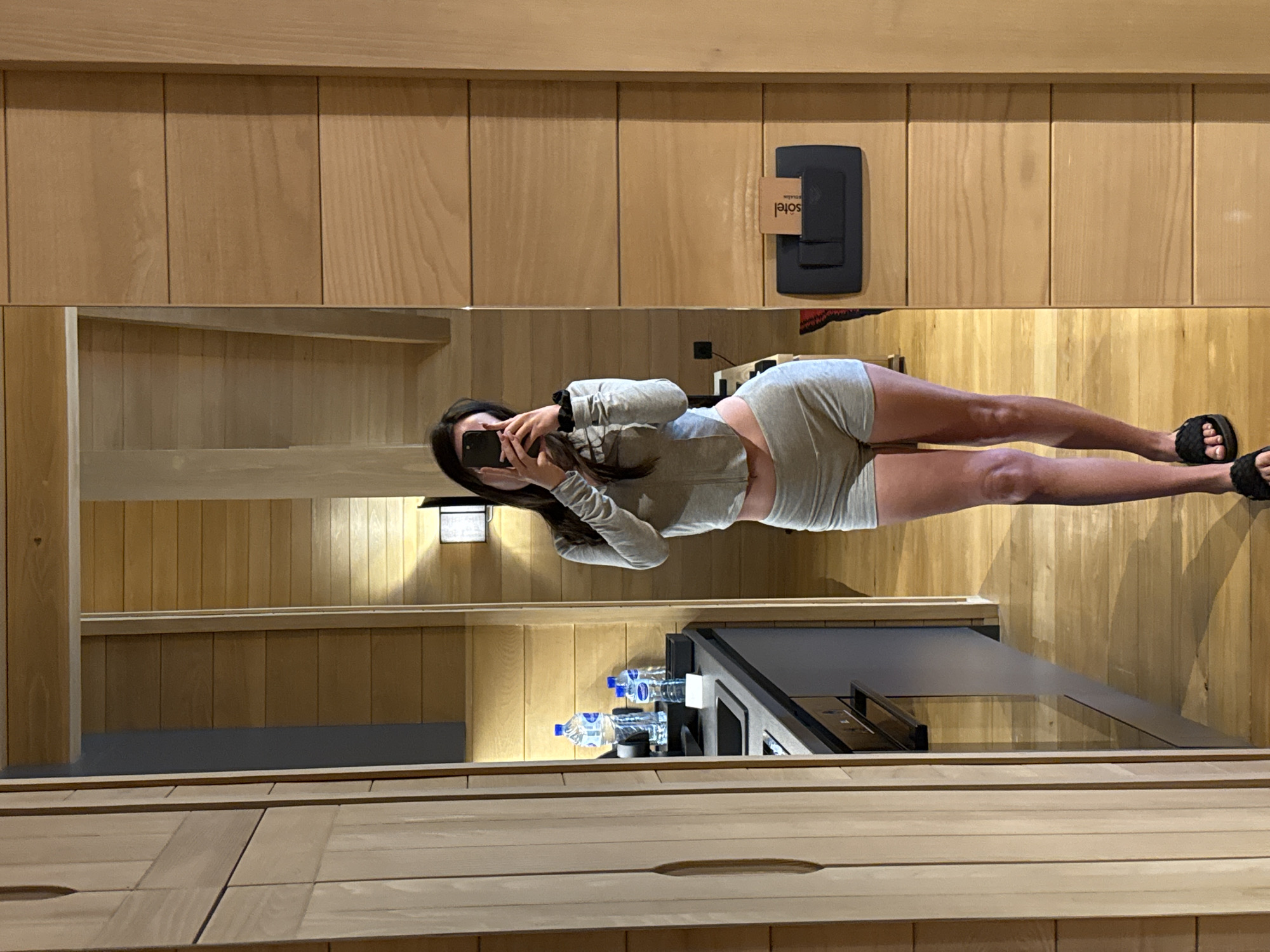 The image shows a young woman taking a mirror selfie in a room with wooden paneling. She is wearing a grey long-sleeved crop top and grey shorts. She is holding a black smartphone to take the picture. The room appears to be a hotel room, as indicated by a "so tel" sign on the wall, which is partially obscured. In the reflection of the mirror, we can see a desk with several bottles of water and a shelf. The lighting suggests it could be daytime, or at least well-lit indoors. The woman's posture and the selfie suggest a casual and perhaps playful moment.