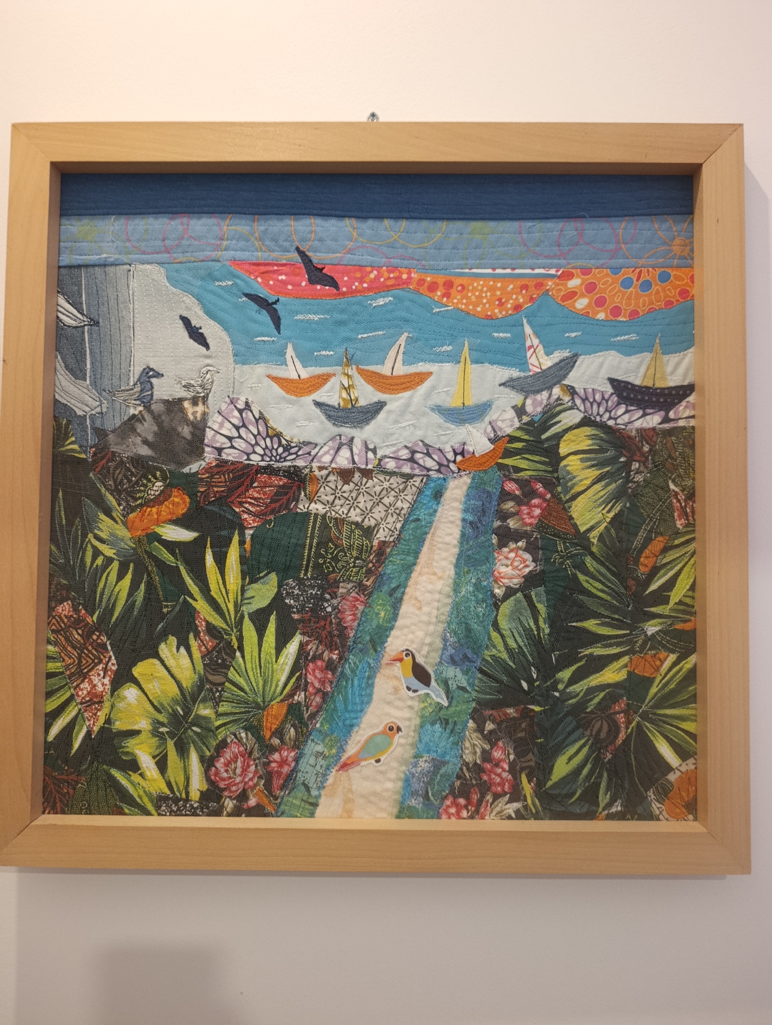 This is a close-up, eye-level shot of a framed fabric collage artwork. The artwork depicts a vibrant, stylized coastal scene. The sky is a bright blue with swirling patterns of orange and pink. A body of water, possibly the sea, is rendered in various shades of blue and white, with several sailboats with orange and yellow sails navigating its surface. Flocks of black birds are scattered across the sky.
The foreground of the artwork is dominated by lush, green tropical foliage, with large leaves and colorful flowers in shades of red and pink. A pathway, possibly leading to the water, is visible in the center, with two small, colorful birds, one with yellow and black markings and another with orange and green, perched on it. The overall impression of the artwork is cheerful and lively, suggesting a sunny day at a tropical beach. The artwork is framed in a simple, light-colored wooden frame and appears to be hung on a white wall. No visible text is present in the image.