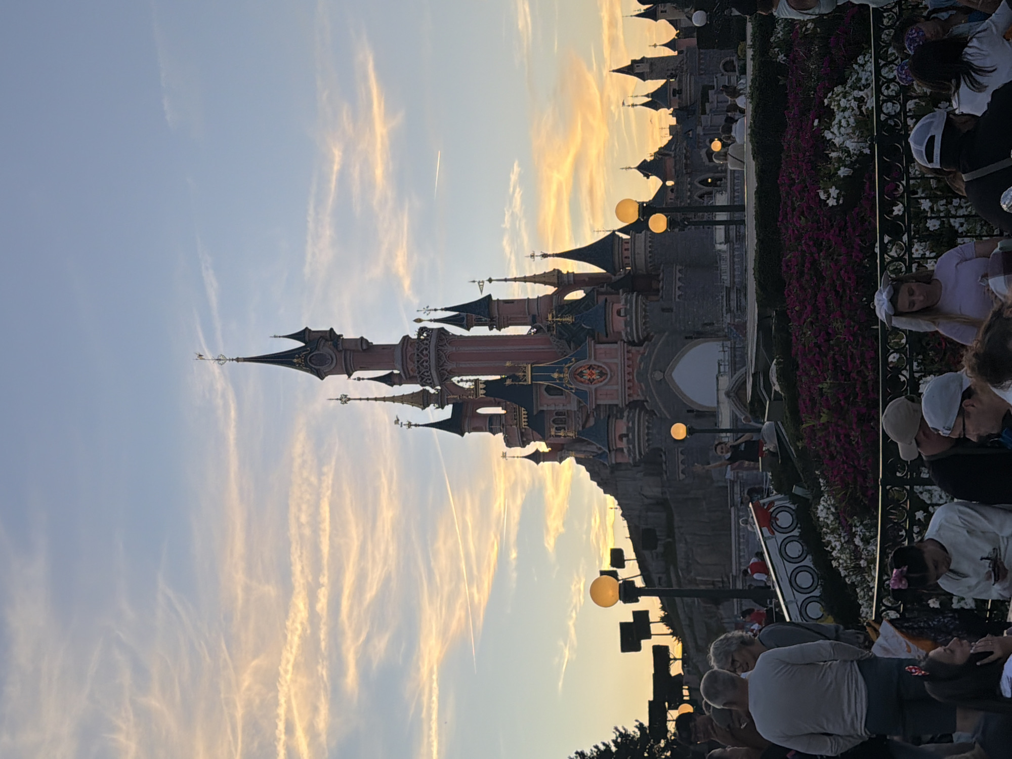 The image shows Sleeping Beauty Castle at Disneyland Paris in Chessy, France, at dusk. The sky is filled with wispy clouds, tinged with orange and yellow from the setting sun, with a contrail visible in the upper portion of the sky. The castle, with its distinctive spires and pink and grey facade, dominates the background. In the foreground, a crowd of people are gathered, some seated and some standing, looking towards the castle or engaged in conversation. Flowerbeds filled with purple and white flowers are visible in the middle ground. Streetlights with glowing orange bulbs are also scattered throughout the scene, illuminating the gathering. The overall atmosphere appears to be one of leisure and observation.