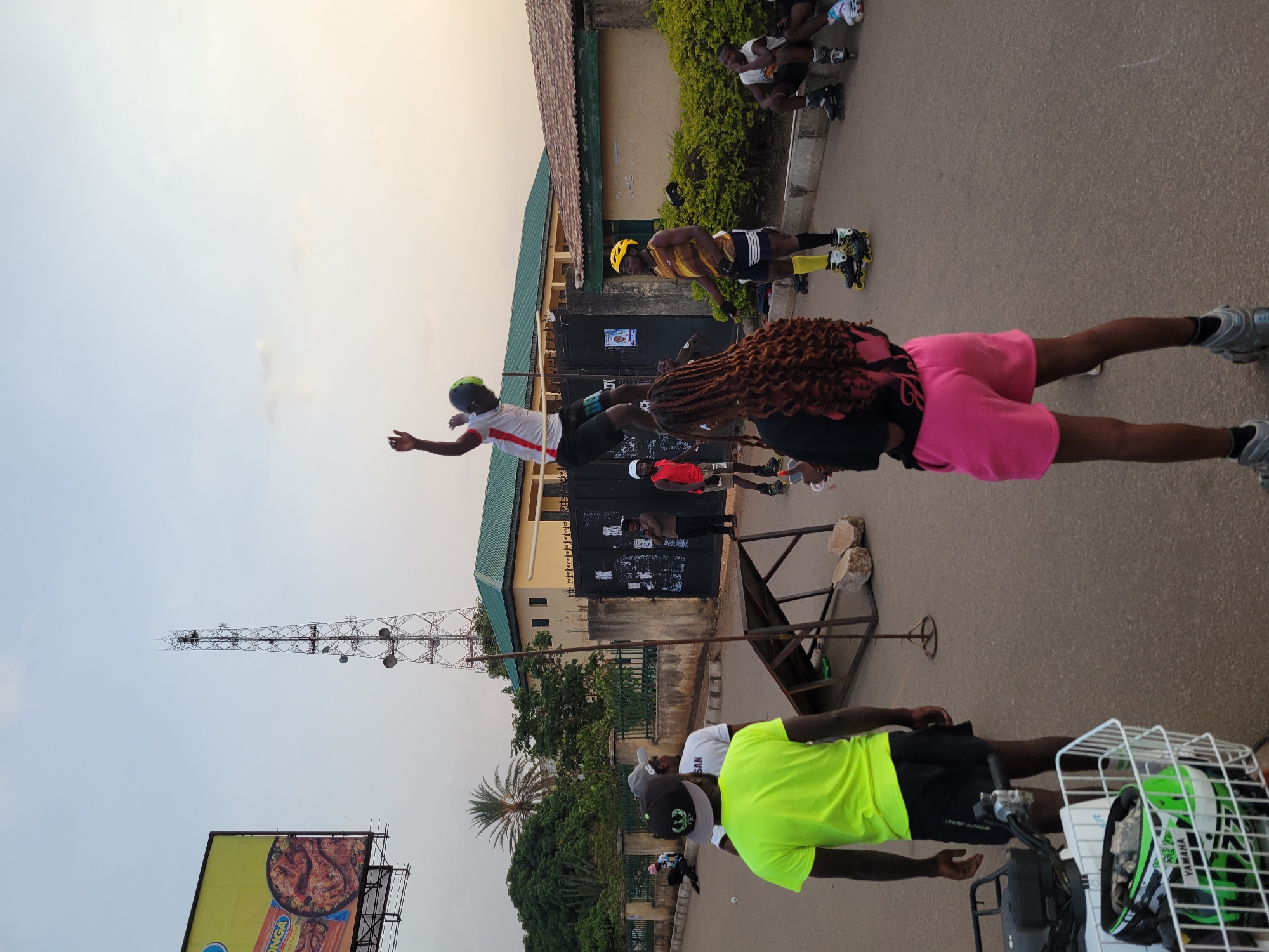 In Jos, Nigeria, a group of people are rollerblading on a street. There is a cell tower and billboard in the background. The group is diverse in age and attire, with some wearing helmets and others not. One person is airborne. There is a barrier in the street, and a scooter parked on the side.