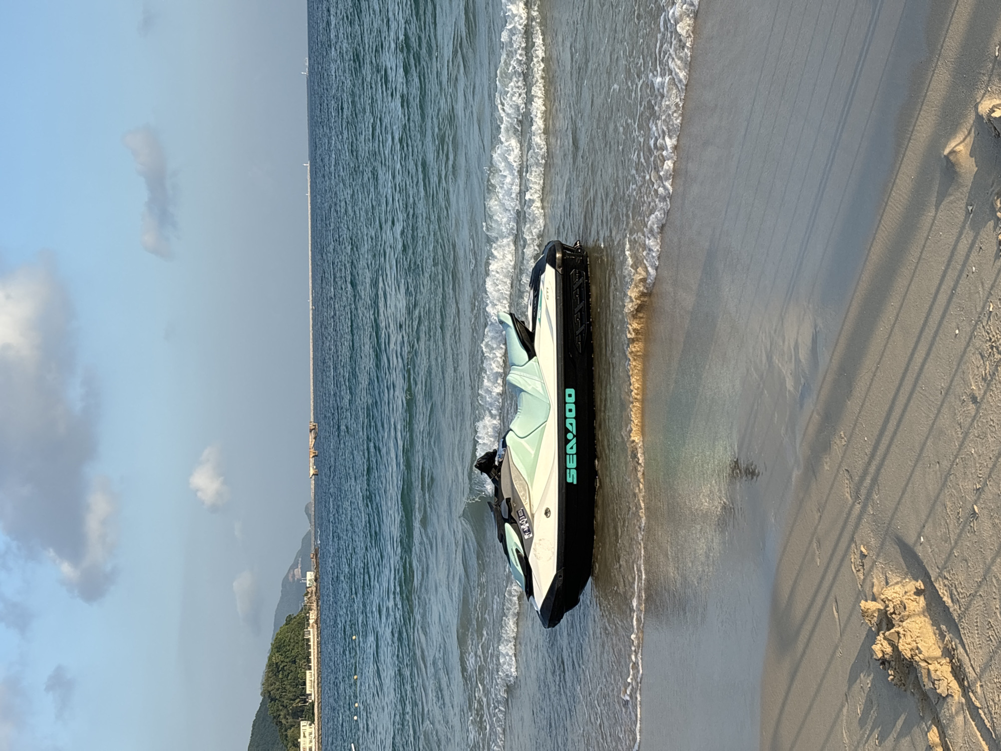 A jet ski, labeled "SEA-DOO," is partially submerged in shallow water at the edge of a sandy beach. The jet ski is white and light blue with black accents. Gentle waves are washing ashore around the jet ski, creating white foam. The beach is light brown sand, with some small piles of sand and shadows cast by an unseen object. In the background, the ocean stretches to the horizon under a light blue sky with scattered clouds. A breakwater, some buildings on a green hill, and a distant shoreline are visible. The overall scene depicts a calm beach environment.