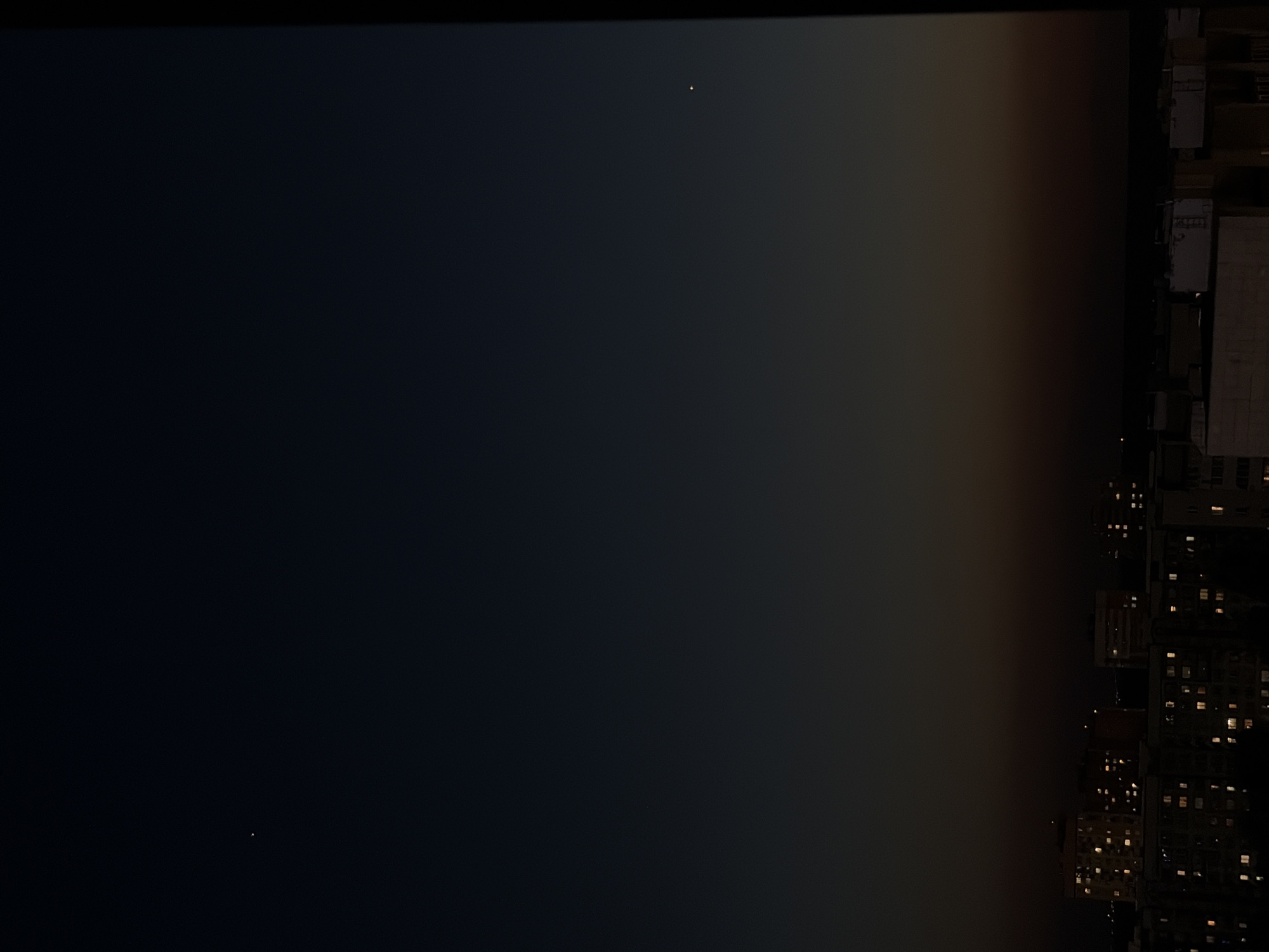 The image depicts a night scene, likely an aerial view, showing a dark sky and the illuminated skyline of a city. The right side of the frame features buildings with lit windows, suggesting residential or office spaces. A gradient of light transitions from dark sky to the city's glow, indicating the presence of atmospheric light or pollution. Two small points of light, likely stars or distant city lights, are visible against the dark sky. The city is Naberezhnye Chelny, Russia.