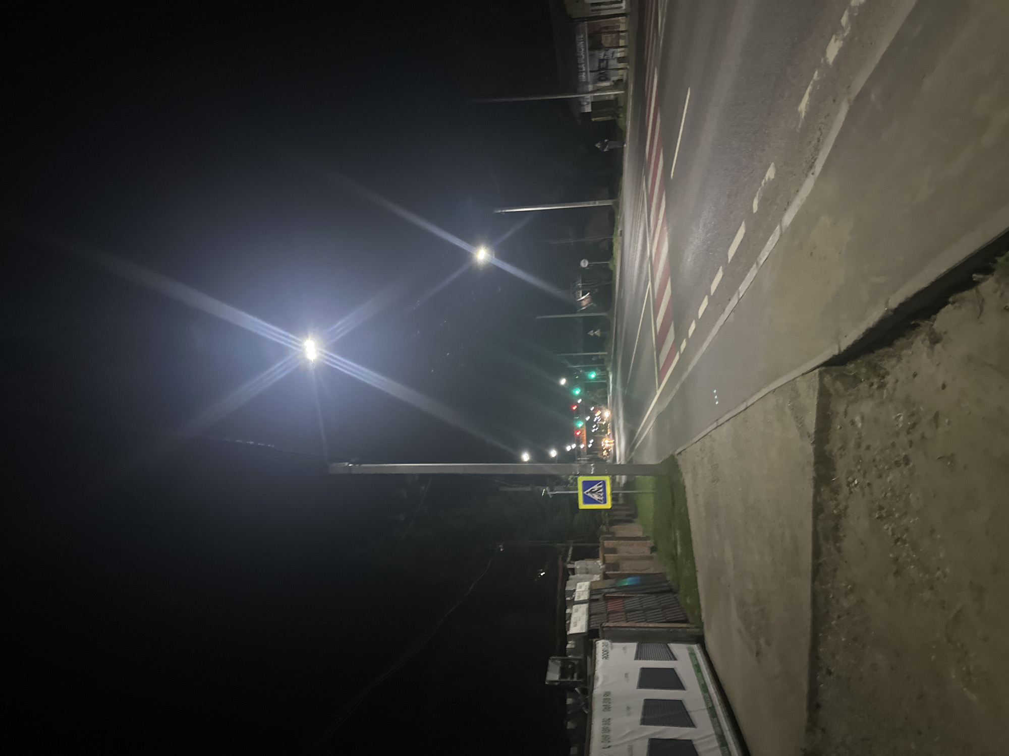 The image captures a street scene at night in the city of Strășeni, Moldova. A pedestrian crossing with red and white markings is visible on the asphalt road. Streetlights illuminate the area, casting bright beams and creating starburst effects due to the camera's aperture. Along the left side of the road, there is a sidewalk, followed by a grassy area and then a fence. Beyond the fence, there are buildings or structures, and some materials that appear to be stacked or stored. Traffic lights are also visible, with a green light illuminated. A pedestrian crossing sign, a yellow diamond with a black silhouette of a person walking, is present near the sidewalk. In the distance, headlights from vehicles suggest ongoing traffic. The sky is dark, indicating nighttime.
