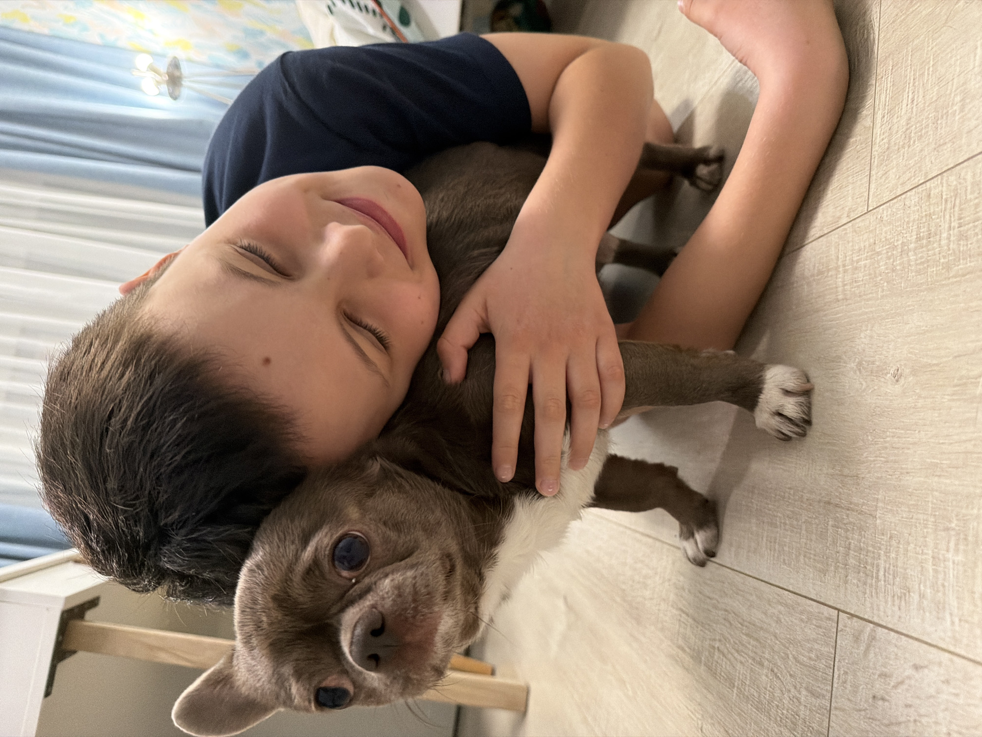 A boy is embracing a dog on a wooden floor. The boy has closed eyes and is smiling. The dog is brown and white. The location is a room with wooden floors. The city of Votkinsk, Russia is not discernible from the image.