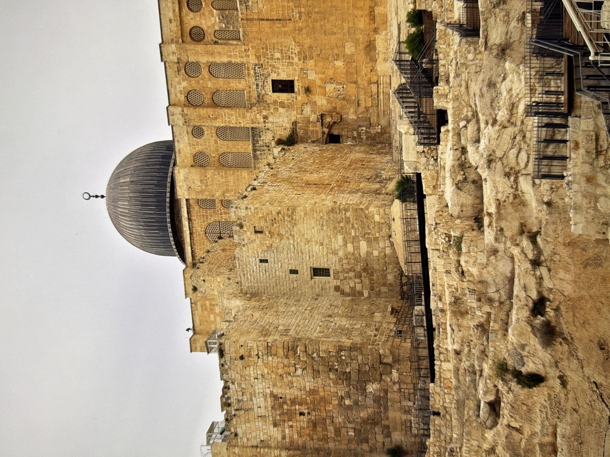 The image shows a view of the Al-Aqsa Mosque in Jerusalem, Israel. The mosque is a large stone building with a prominent dark dome. The structure is partially obscured by a rugged, rocky foreground with multiple sets of metal stairs and railings leading up towards the building. There are small green plants growing in various parts of the rocky terrain. The sky above is overcast and light gray. The architectural style of the mosque is characteristic of Islamic religious buildings, with its arched windows and decorative patterns. The stone construction of the walls suggests an ancient origin.
